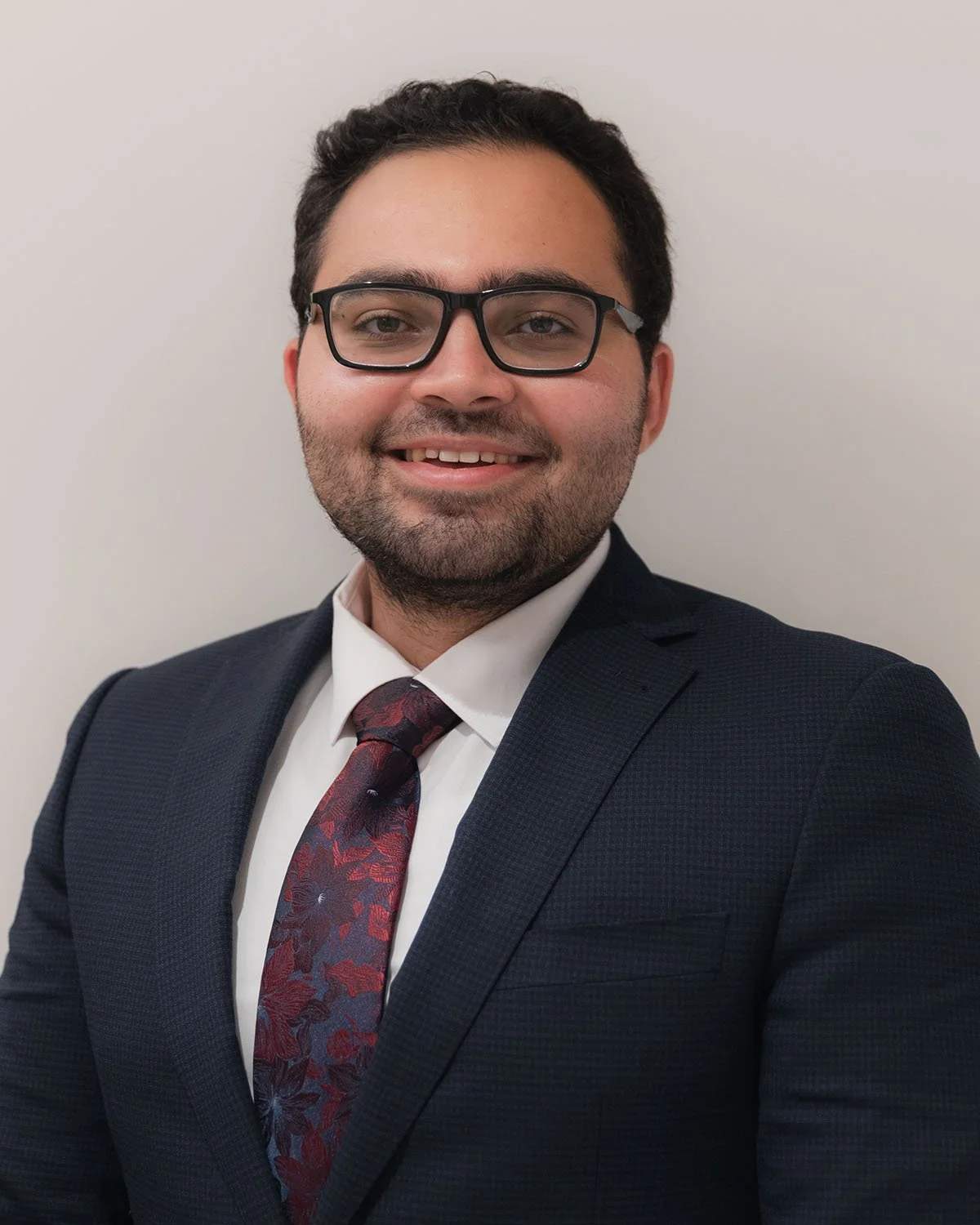 Man smiling in a professional headshot wearing glasses, a dark suit, and a patterned tie against a plain background.