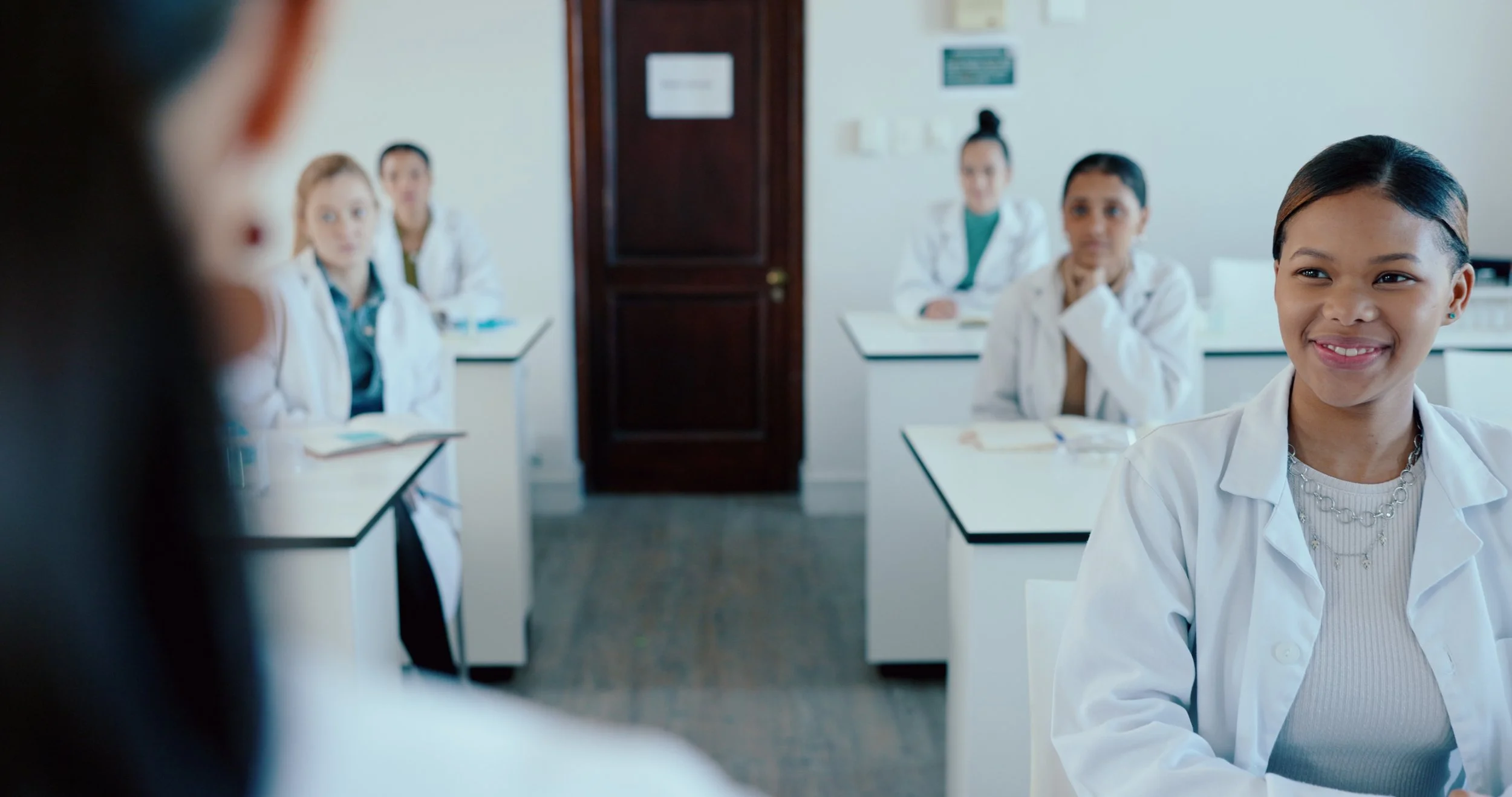 Medical students in white coats sit in a classroom listening to an instructor, with one student in the foreground smiling while others focus on the lesson.