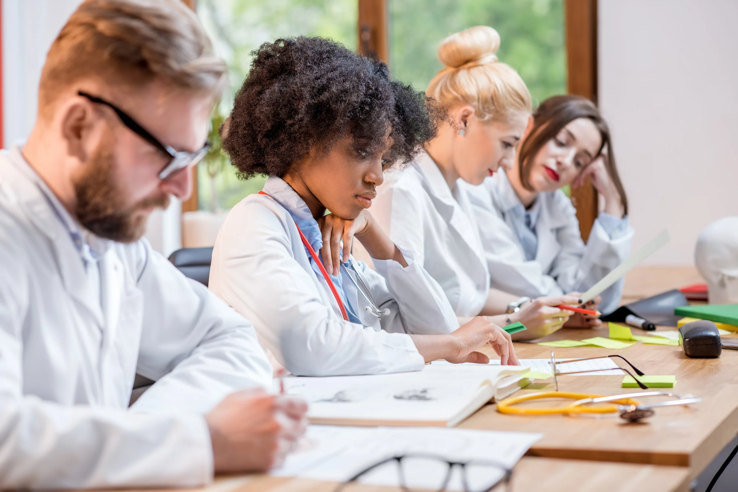 Group of healthcare professionals in white coats sit together at a table, studying notes and reading documents during a training or study session.