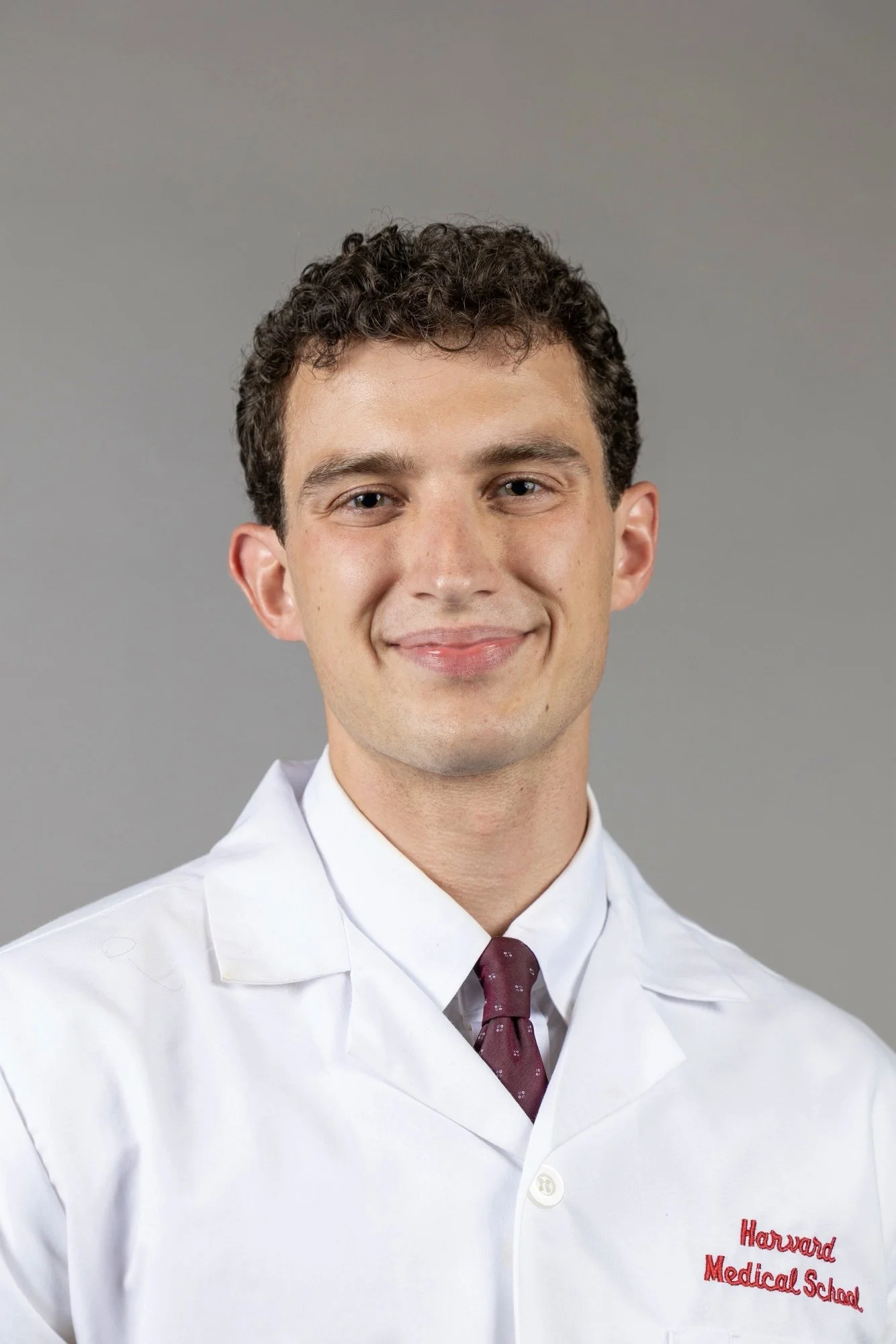 Man smiling in a professional headshot wearing a white medical coat and burgundy tie against a gray background.