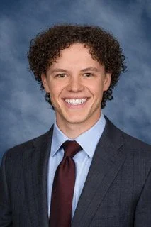 Man smiling in a professional headshot wearing a navy suit, light blue shirt, and burgundy tie against a blue studio background.