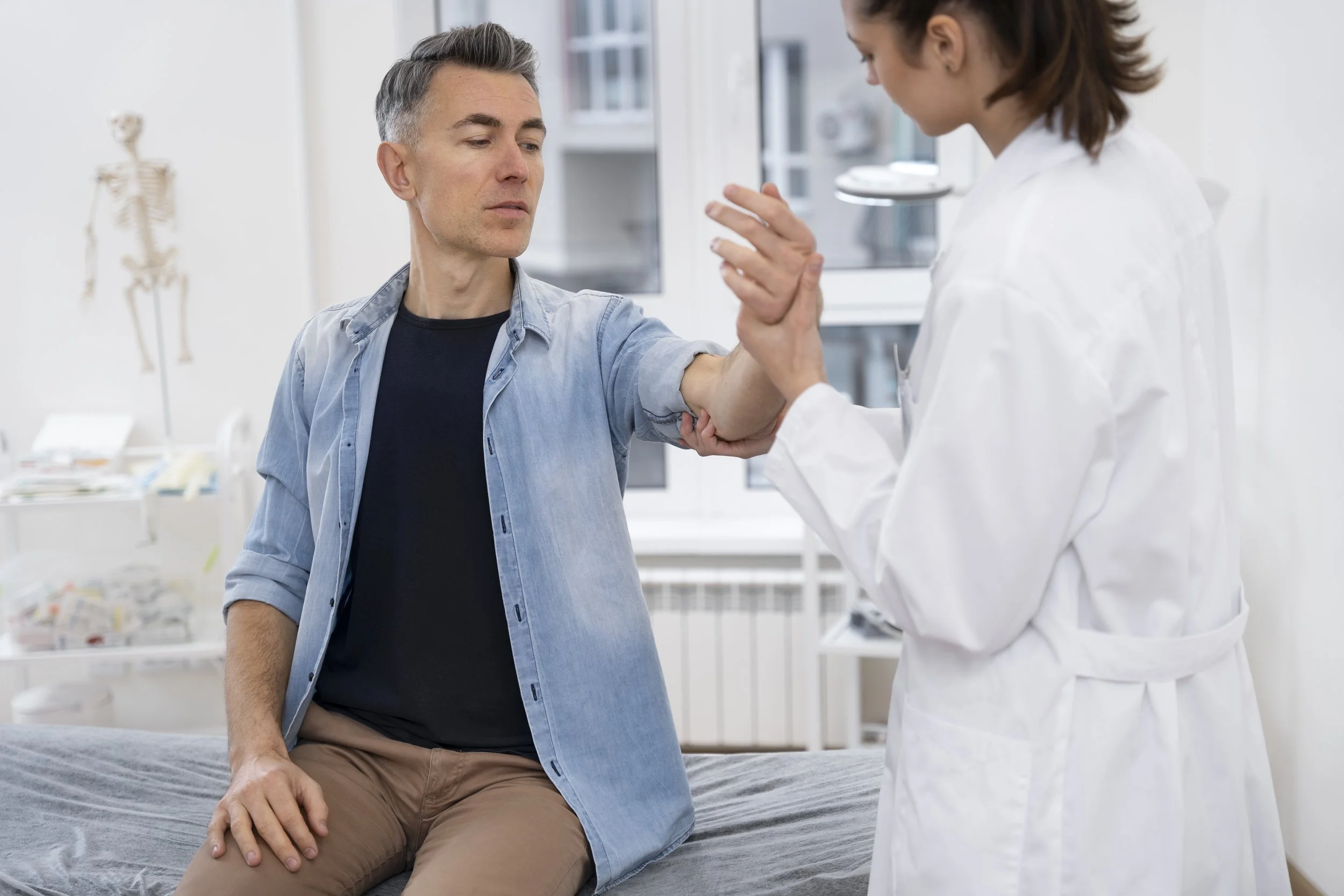 A healthcare professional examines a patient's arm and elbow during a physical assessment, checking range of motion in a bright medical examination room.