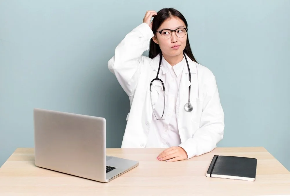 Doctor in a white coat looking confused while sitting at a desk with a laptop and notebook.