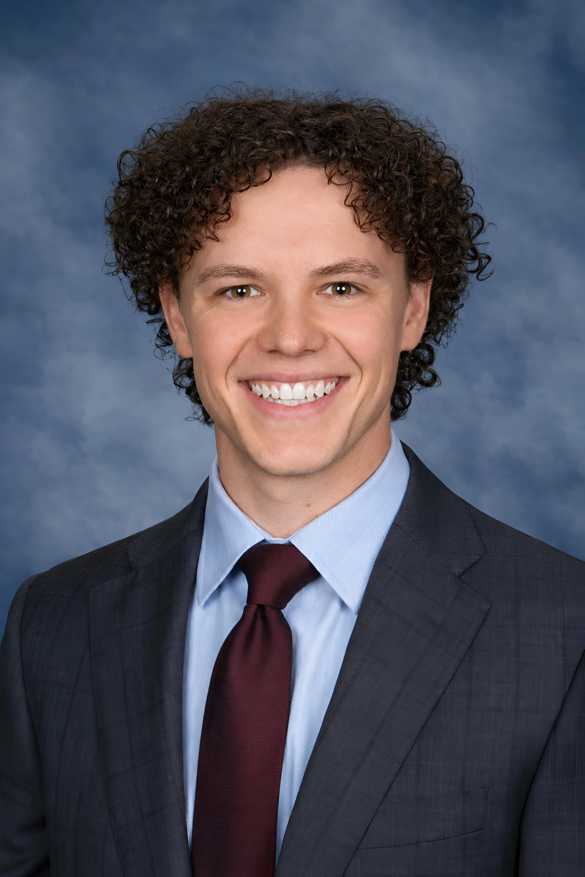 Man smiling in a professional headshot wearing a navy suit, light blue shirt, and burgundy tie against a blue studio background.