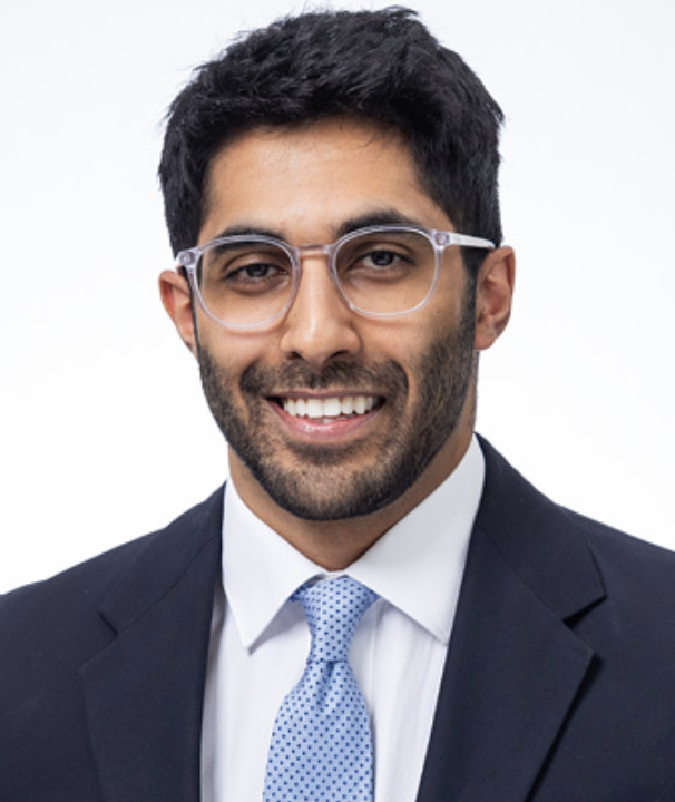 Man smiling in a professional headshot wearing glasses, a navy suit, and a light blue tie against a plain white background.