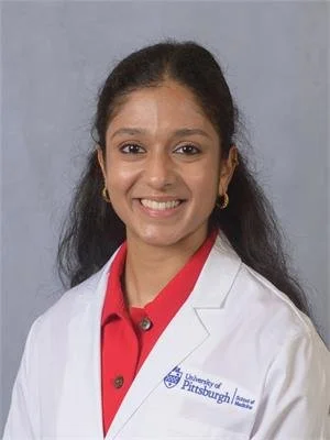 Woman smiling in a professional headshot wearing a white medical coat and red shirt against a gray background.