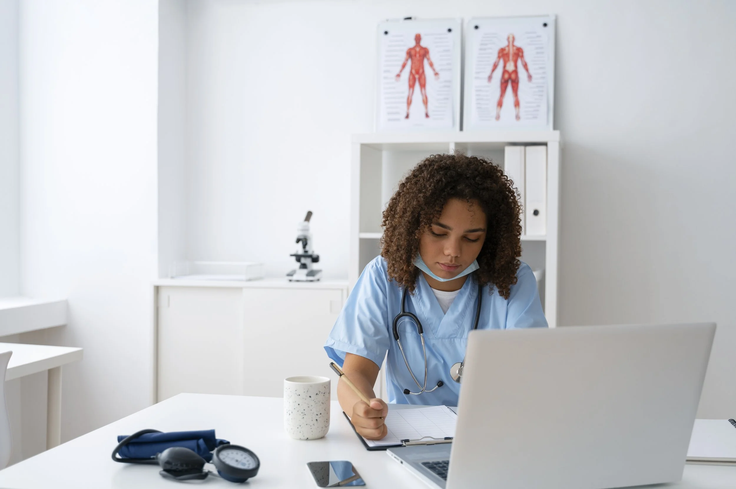 Medical student in scrubs studying at a desk with a laptop and taking notes in a notebook.