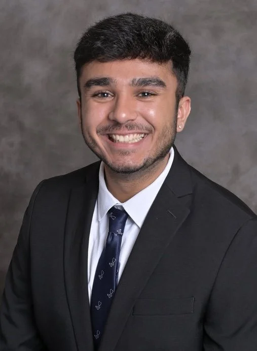 Man smiling in a professional headshot wearing a dark suit and tie against a gray background.