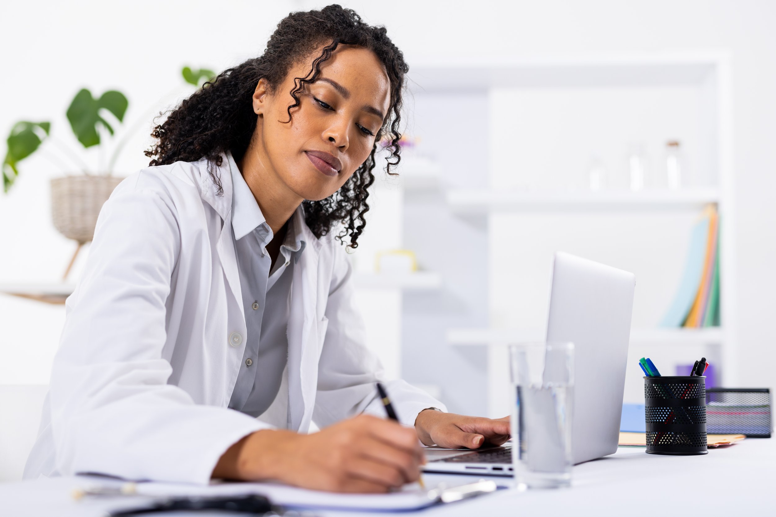A doctor in a white coat works at a desk, writing notes while using a laptop, with office supplies and a glass of water in a bright, modern workspace.