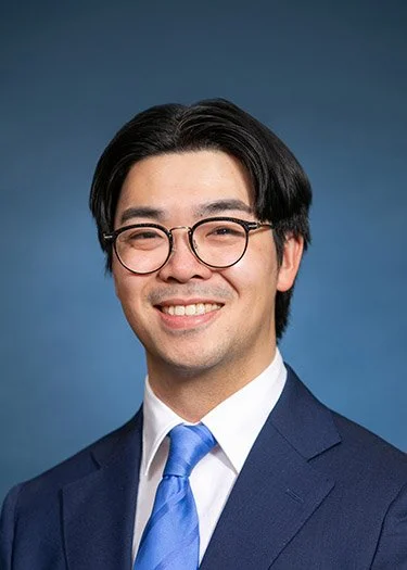 Man smiling in a professional headshot wearing glasses, a navy suit, and a blue tie against a blue background.