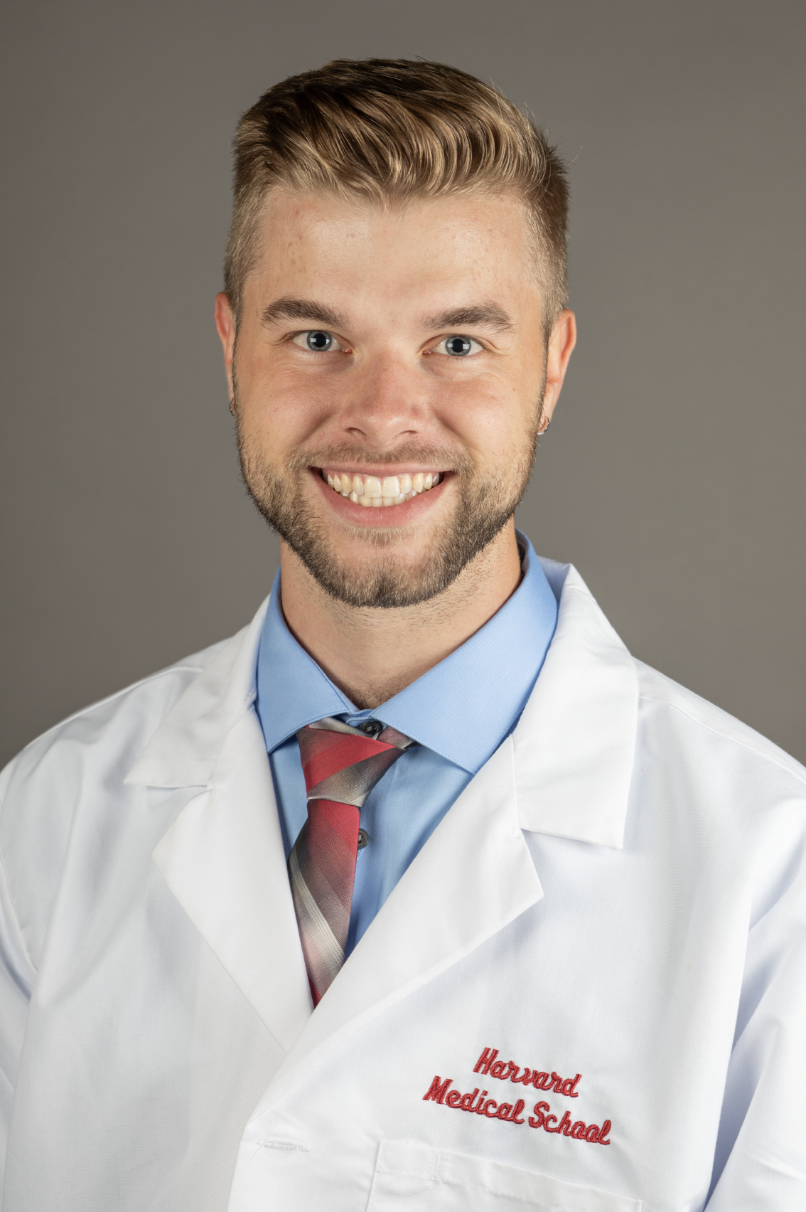 Man smiling in a professional headshot wearing a white medical coat, blue shirt, and striped tie against a gray background.