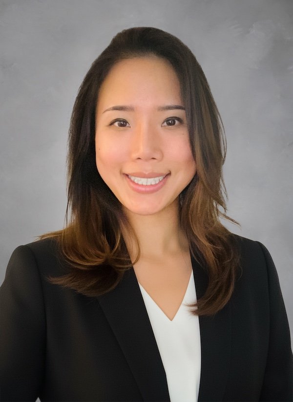 Woman smiling in a professional headshot wearing a black blazer and white top against a gray background.