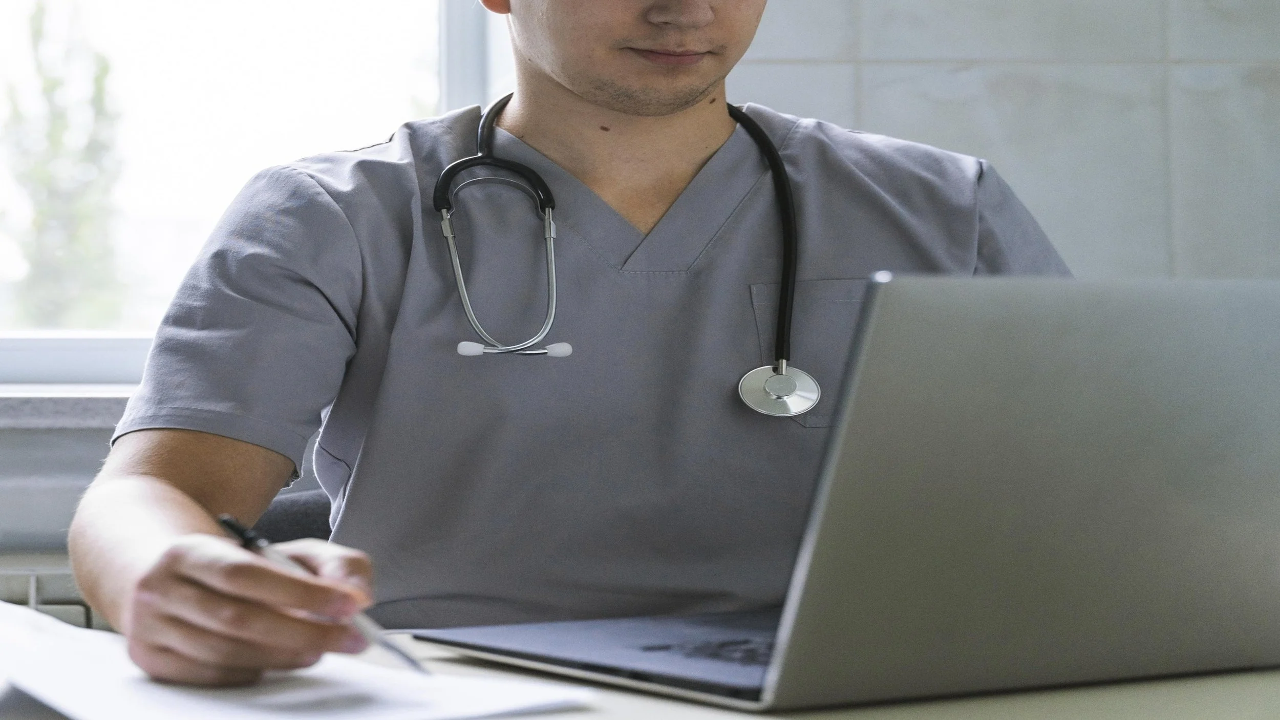 Medical student in scrubs using a laptop and writing notes at a desk.