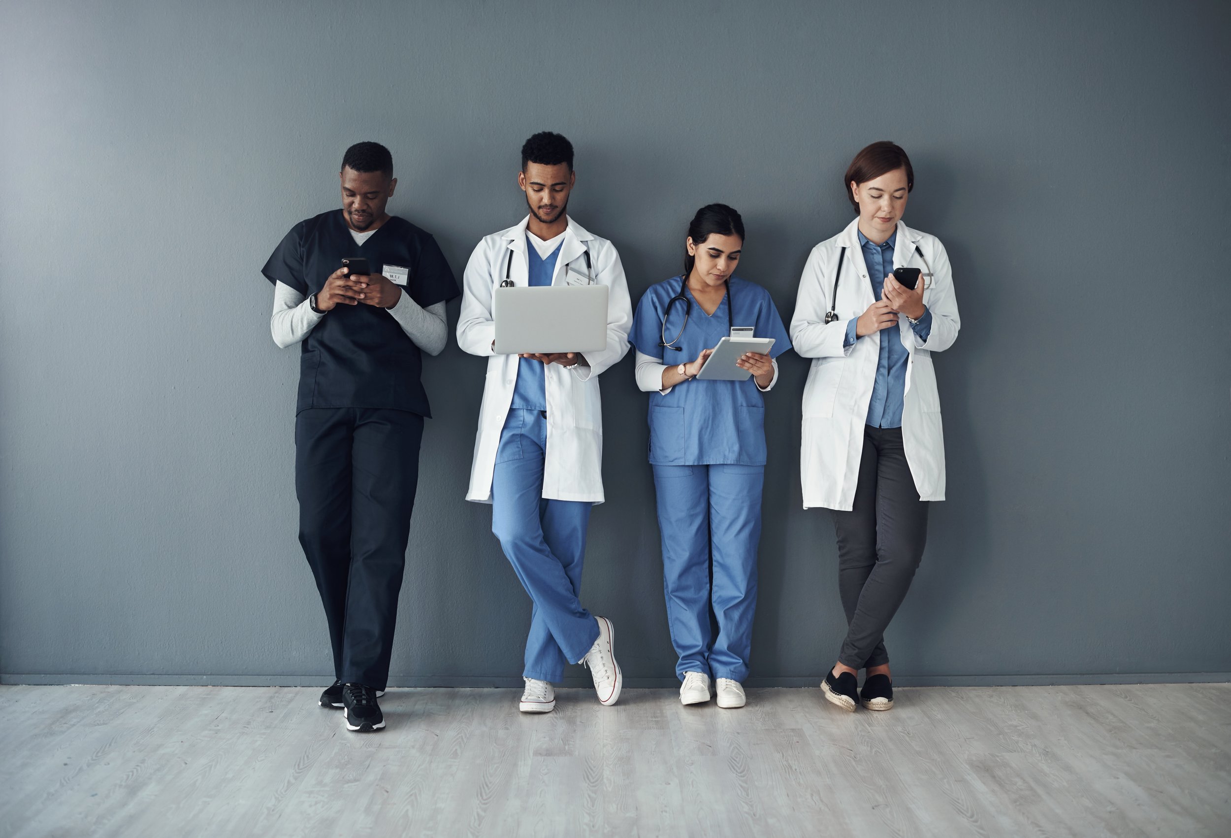 Four healthcare professionals in scrubs and white coats stand against a wall, each using a device—two on smartphones, one on a laptop, and one on a tablet—in a modern clinical setting.