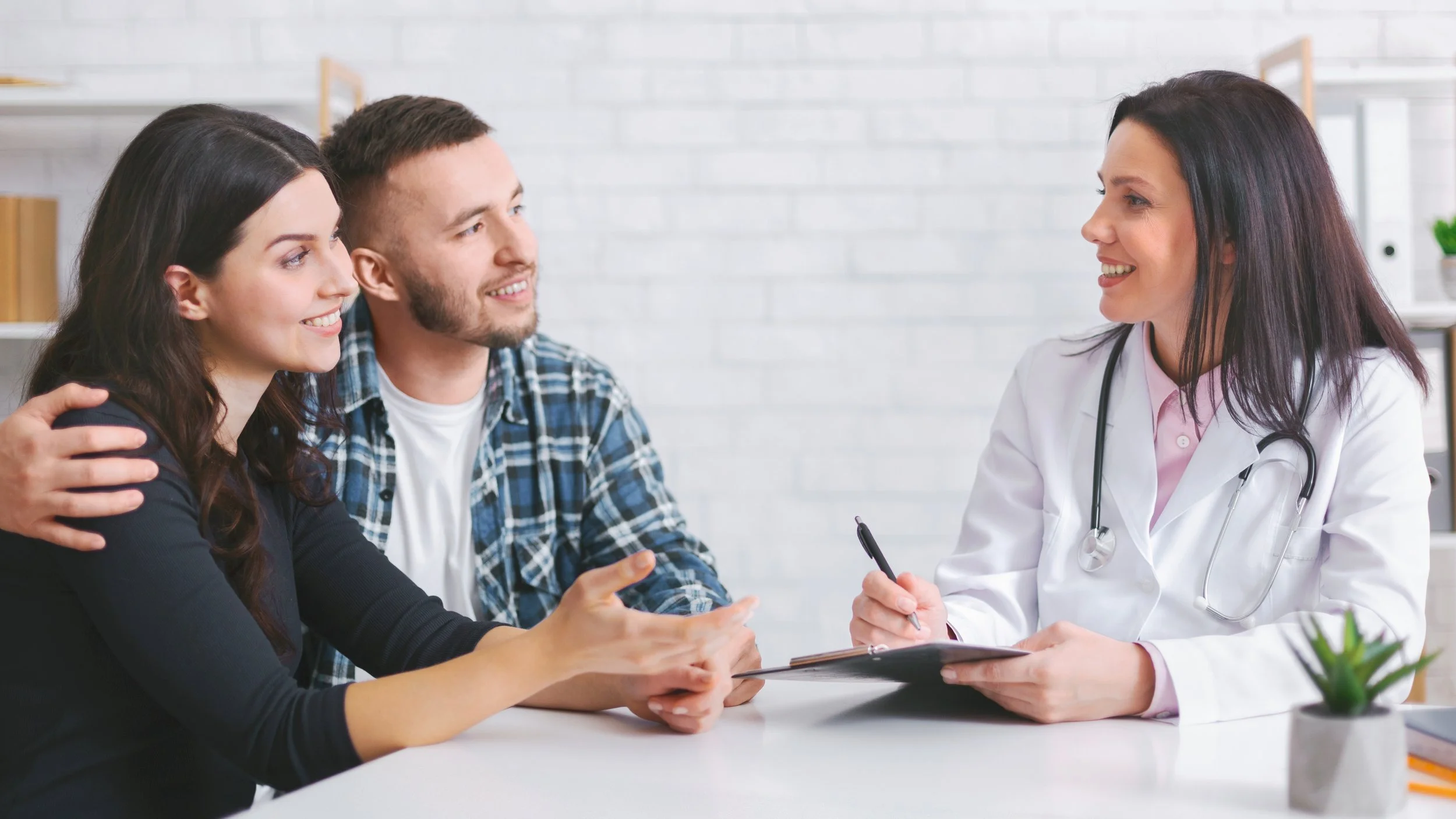 A doctor in a white coat sits at a desk speaking with a smiling couple during a medical consultation, holding a clipboard while the couple listens attentively in a bright clinic office.