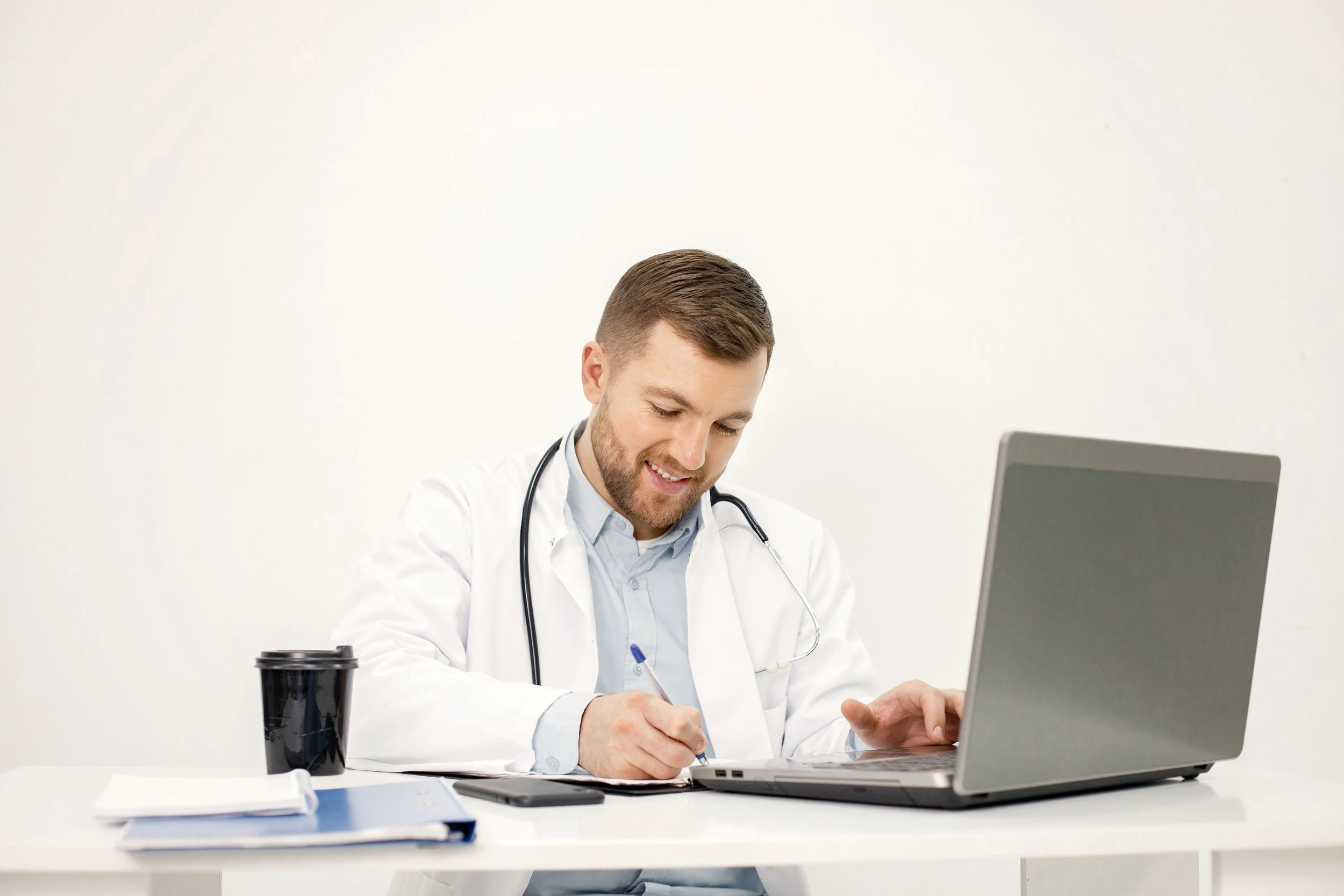 Doctor in a white coat studying and taking notes beside a laptop at a desk.