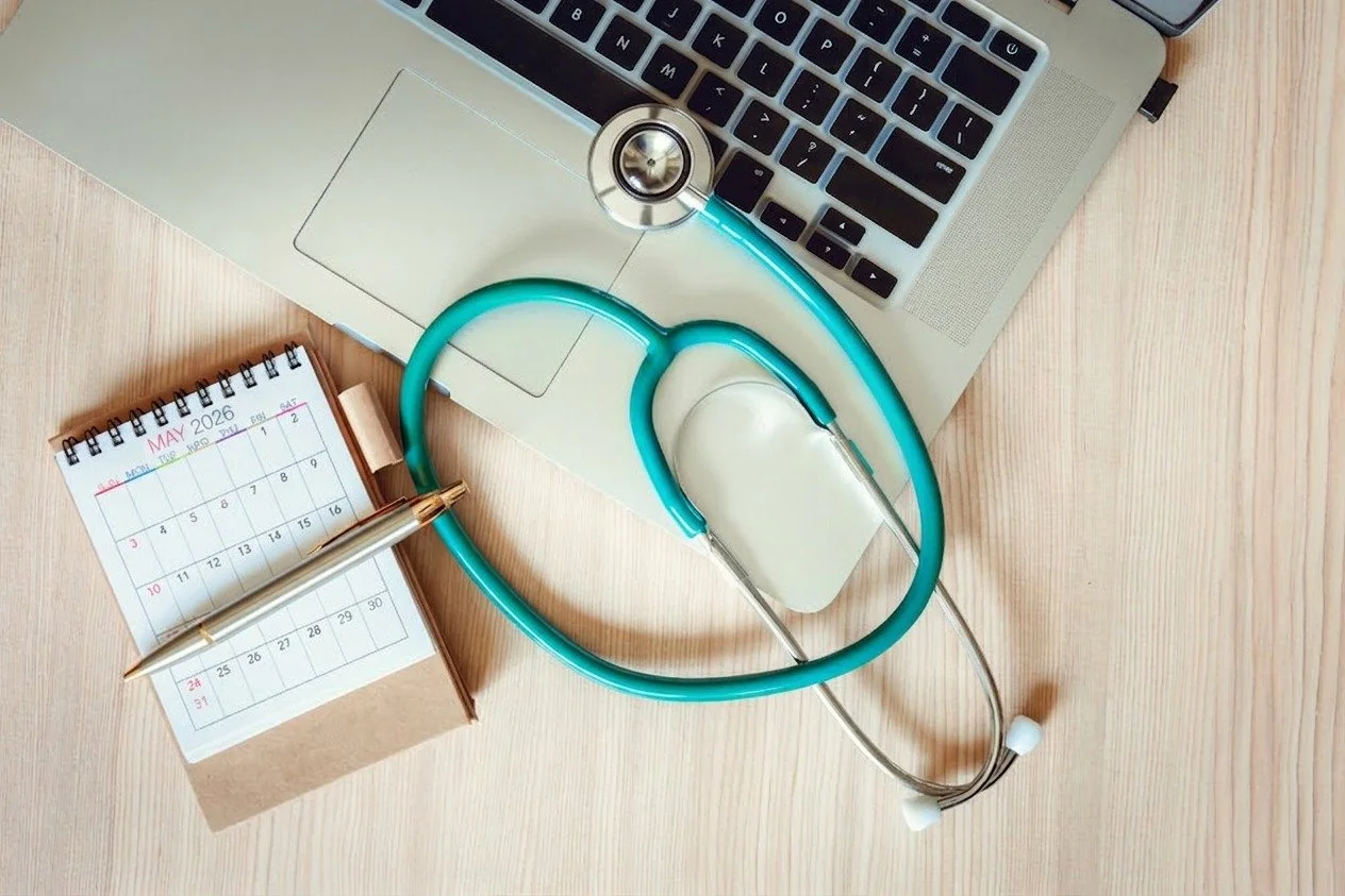 Top-down view of a stethoscope resting on a laptop beside a calendar and pen on a wooden desk, suggesting medical scheduling and work planning.