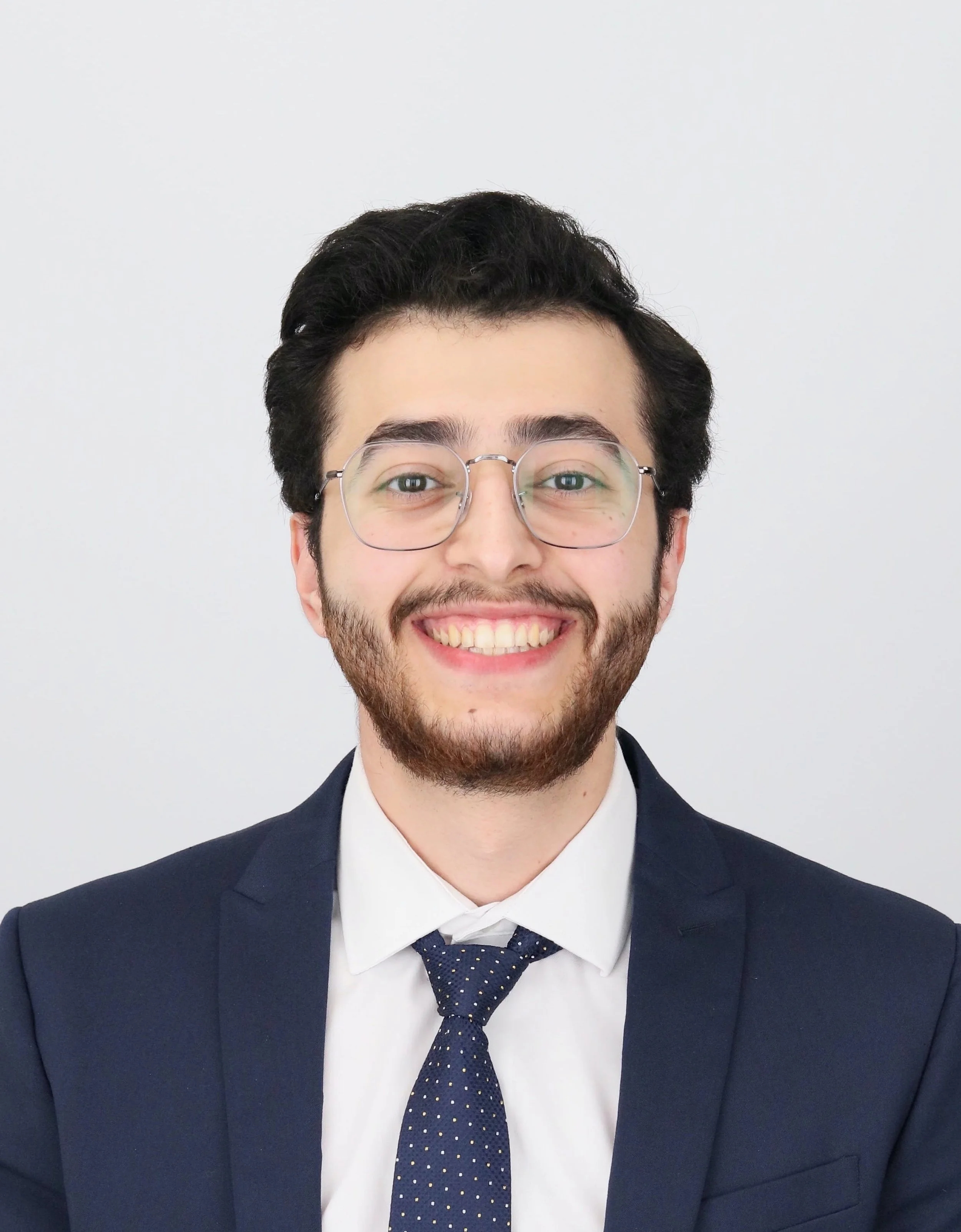 Man smiling in a professional headshot wearing glasses, a navy suit, white shirt, and navy tie against a light gray background.