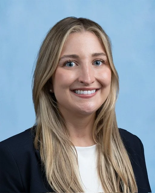 Portrait of a woman with long blonde hair, smiling, wearing a navy blazer and white top, against a light blue background.