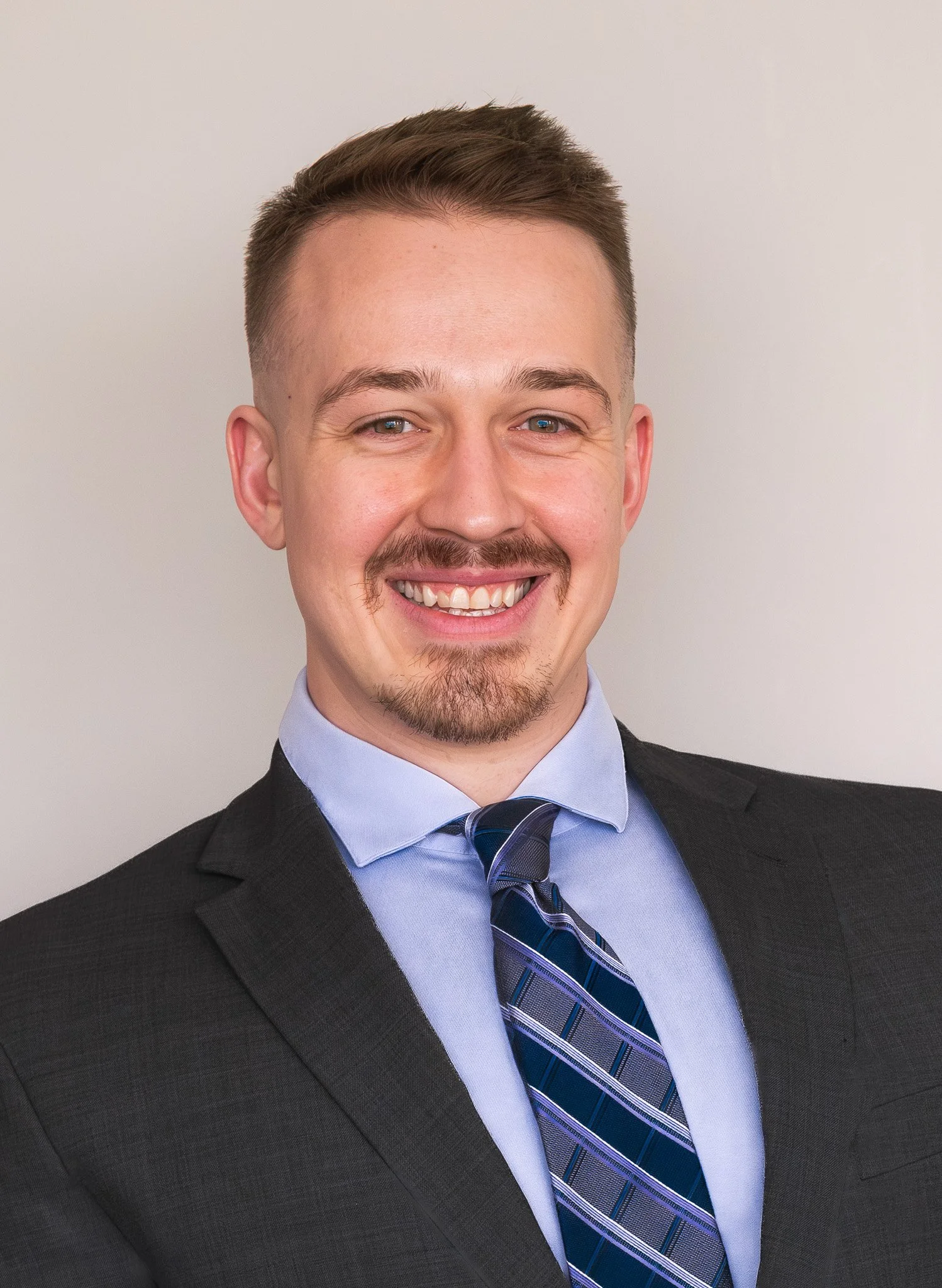 Man smiling in a professional headshot wearing a dark suit, light blue shirt, and striped blue tie against a light gray background.