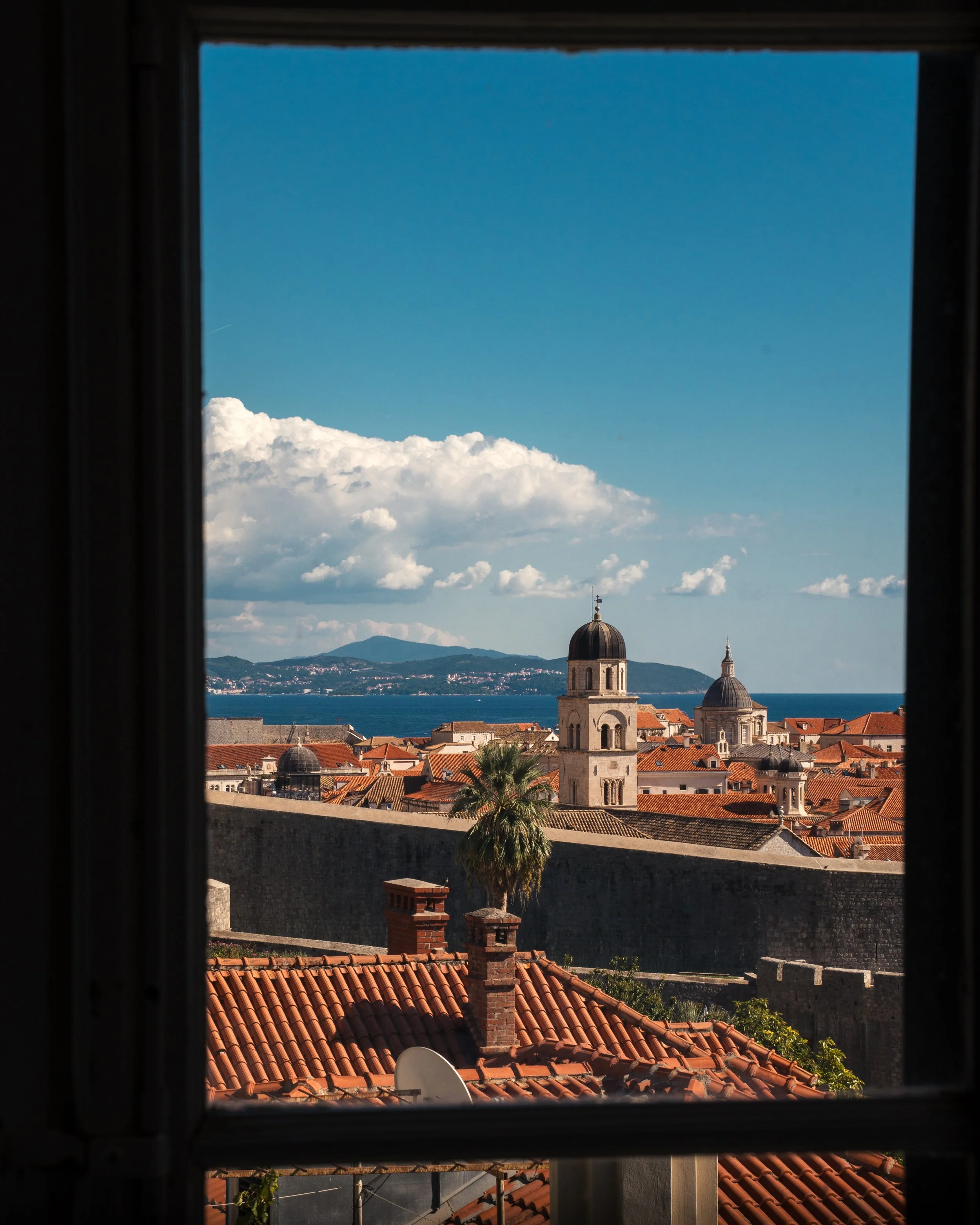 View of a historic European city with orange rooftops and church towers, seen through a window with dark framing, with a distant body of water and hills in the background.