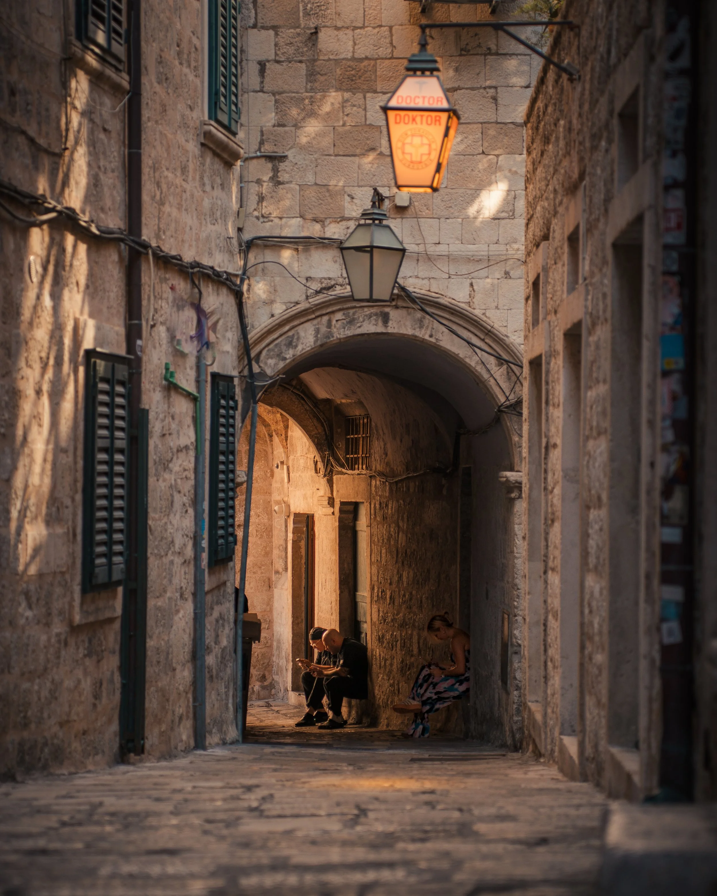 Sunlit narrow cobblestone alleyway with two people sitting on benches, one looking at a phone and the other reading, under an archway with old stone walls and a hanging lantern labeled 'Doctor'.