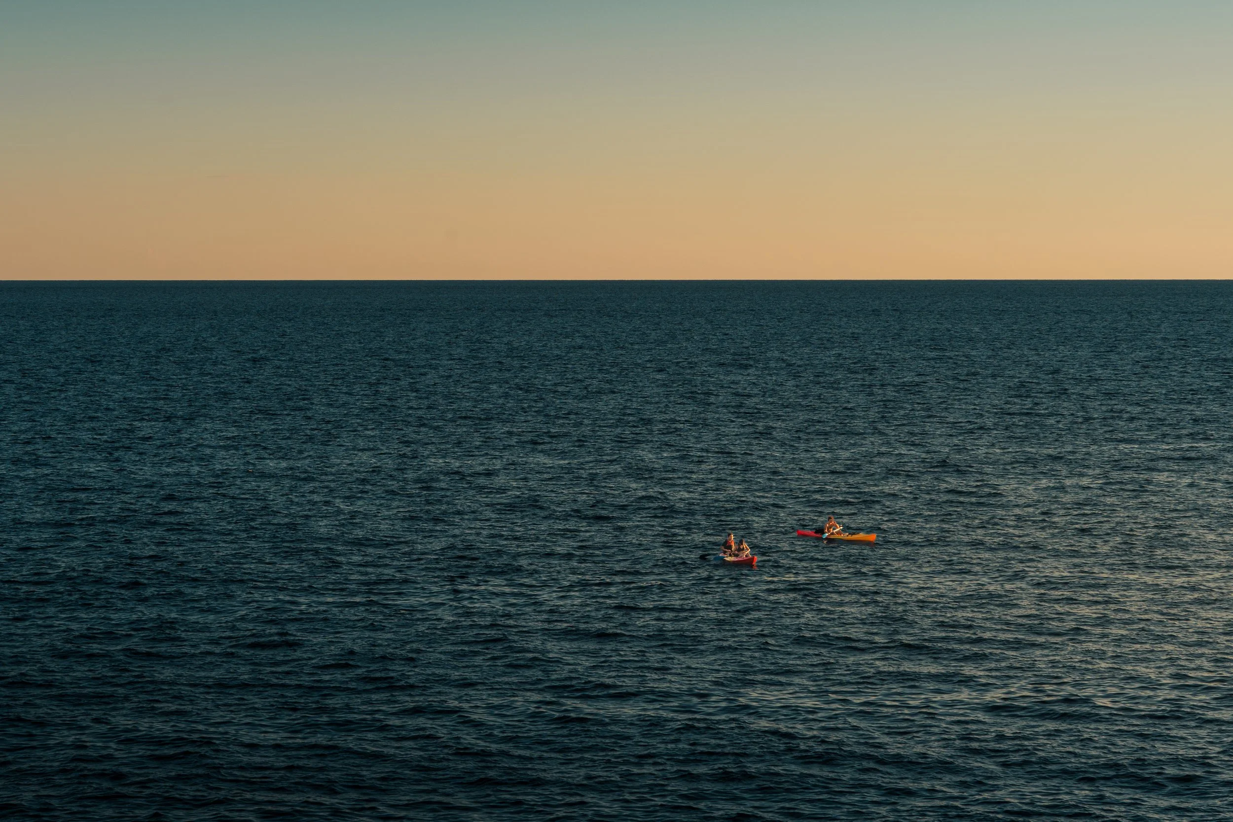 Two people kayaking in the ocean during sunset with a calm water surface and a clear sky.