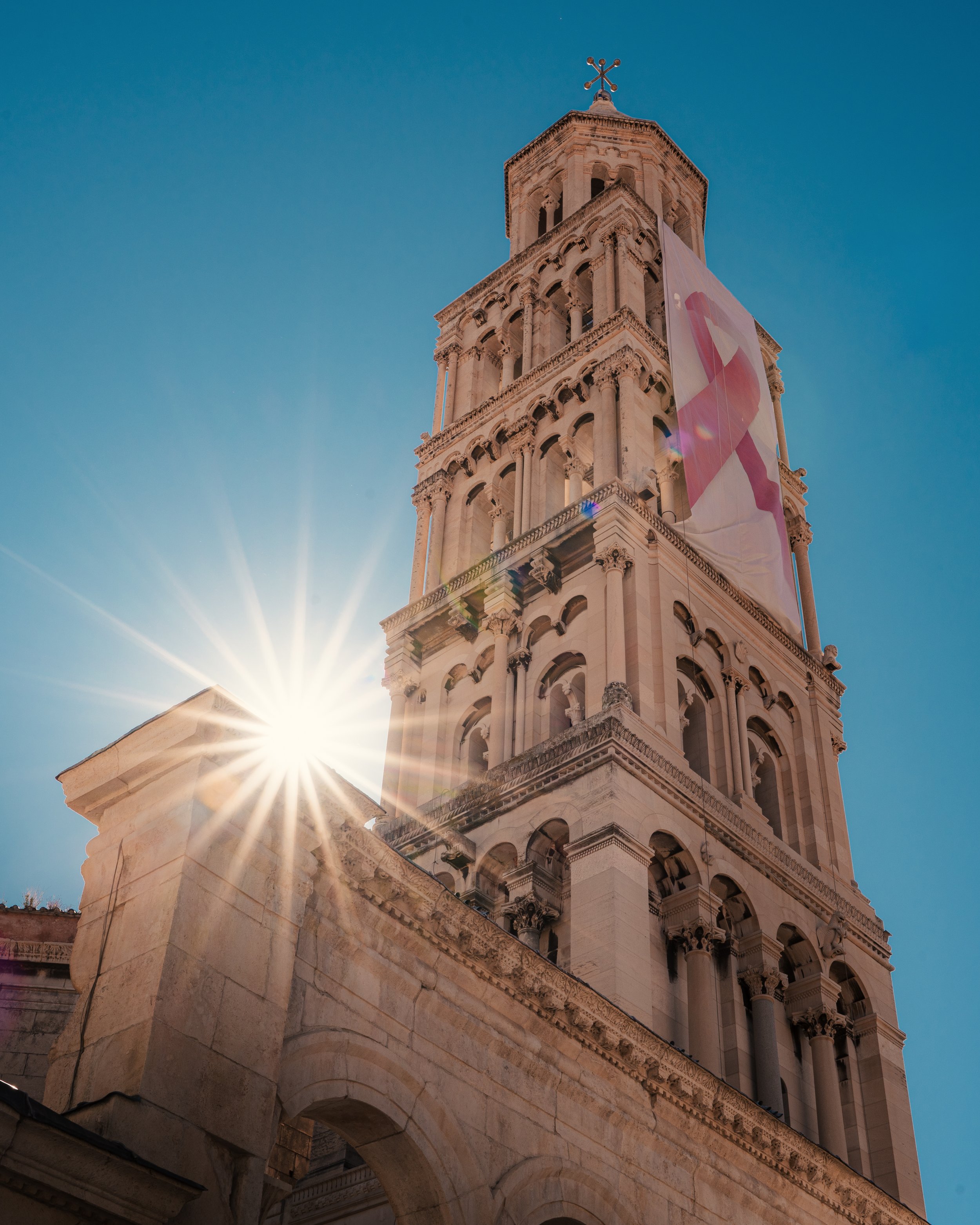 A tall stone bell tower with a cross at the top, under a clear blue sky, with sunlight shining from behind and a pink ribbon banner hanging on its side.
