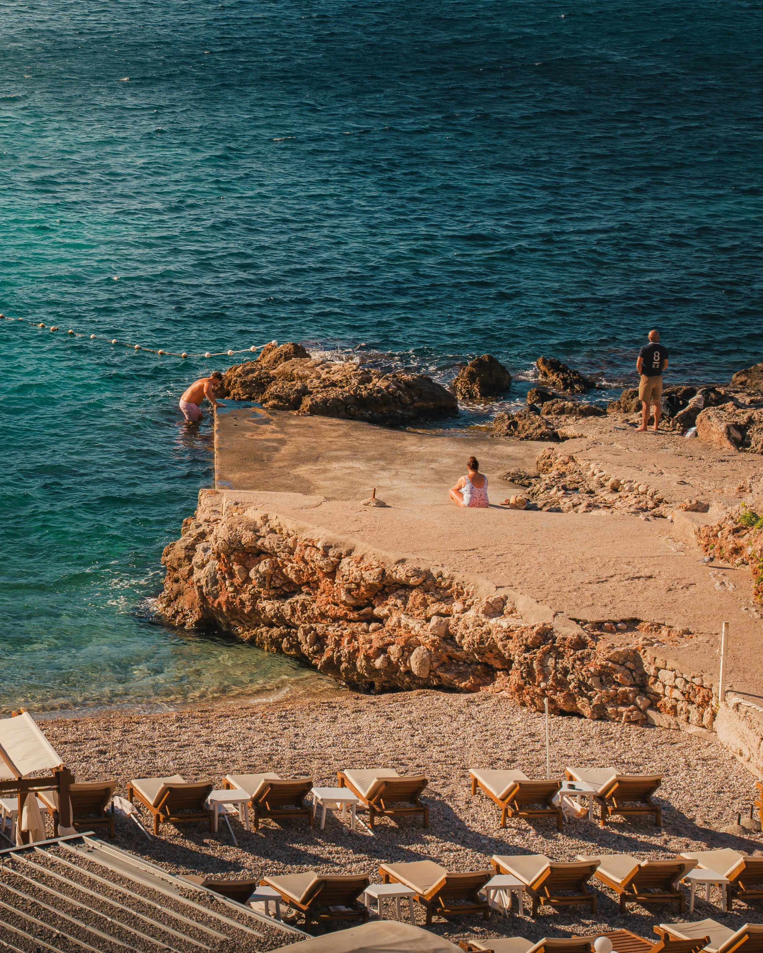 A coastal scene with a concrete pier extending into the ocean, surrounded by rocks. Four people are visible; one is sitting on the pier, two are standing, and one is wading in the water. Beach chairs are lined up on a gravel beach in the foreground.