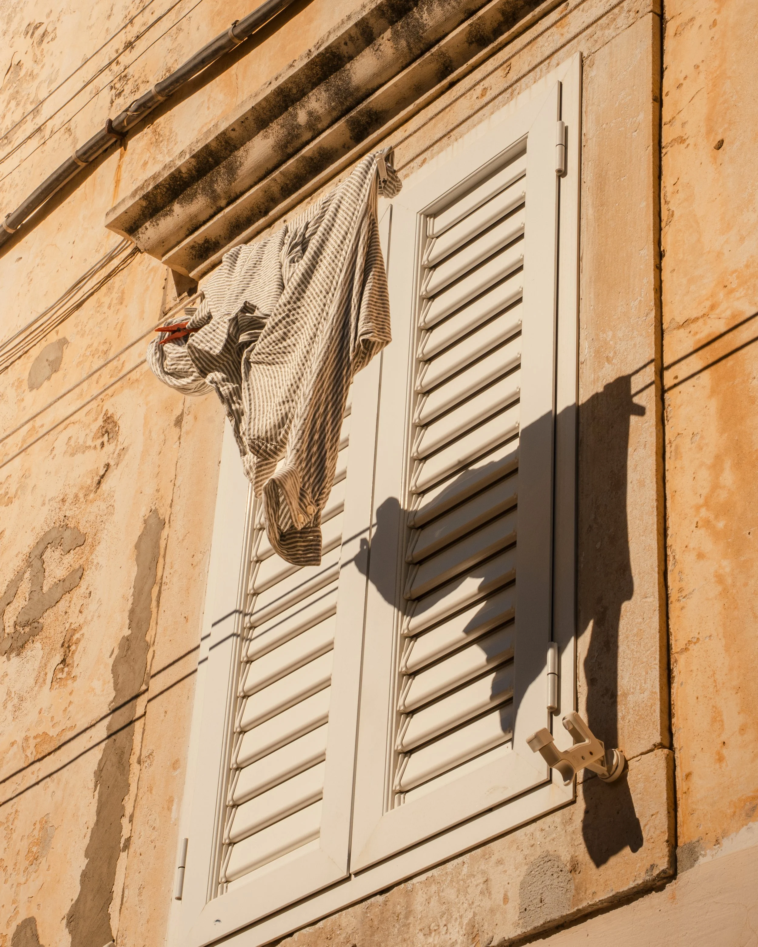 Clothes drying on a clothesline outside a window with closed white shutters, with shadows cast by the sunlight on the wall.