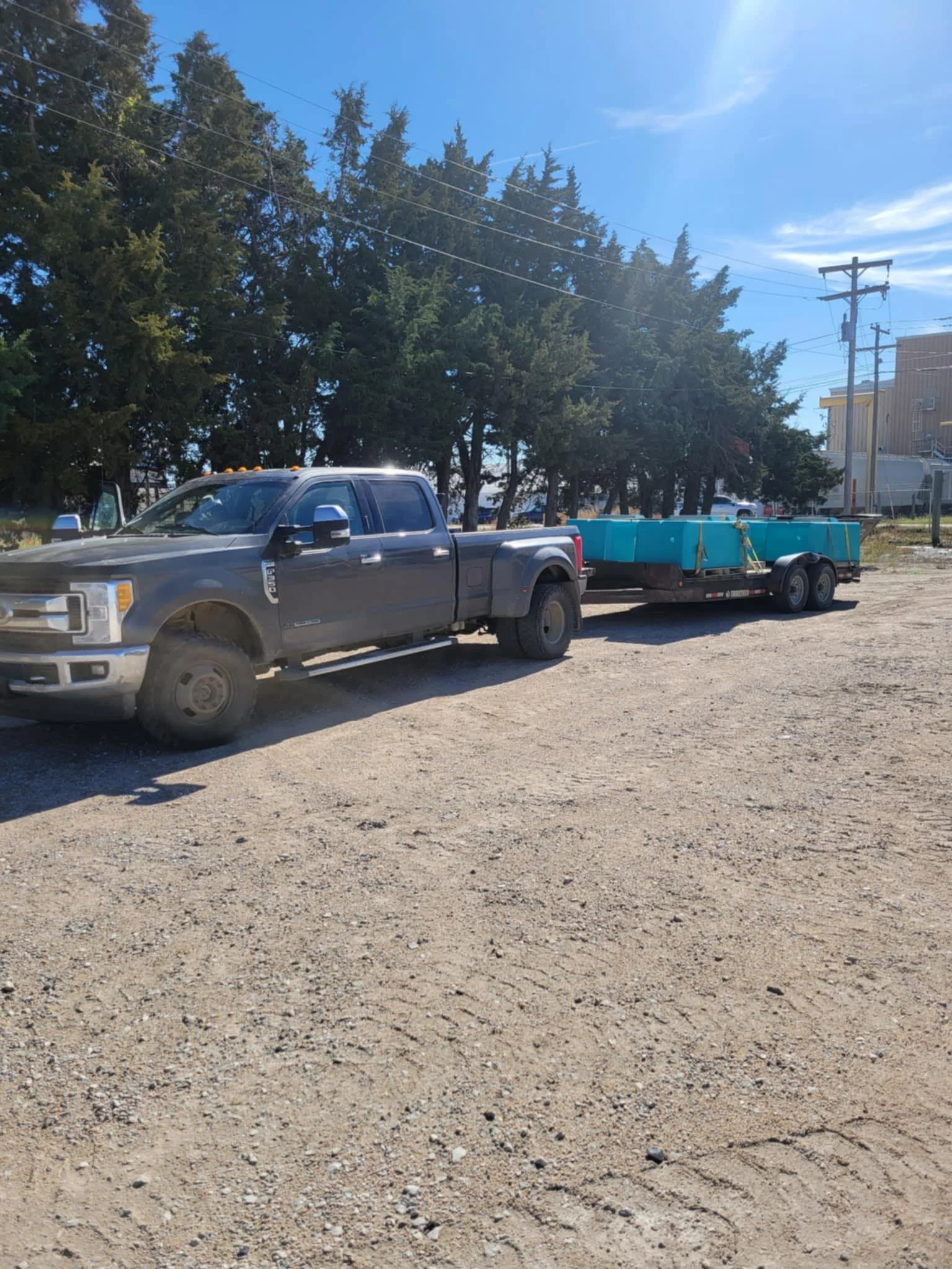 A dark grey pickup truck towing a trailer loaded with Cancrete waterbowls on a dirt lot, with trees and utility poles in the background.