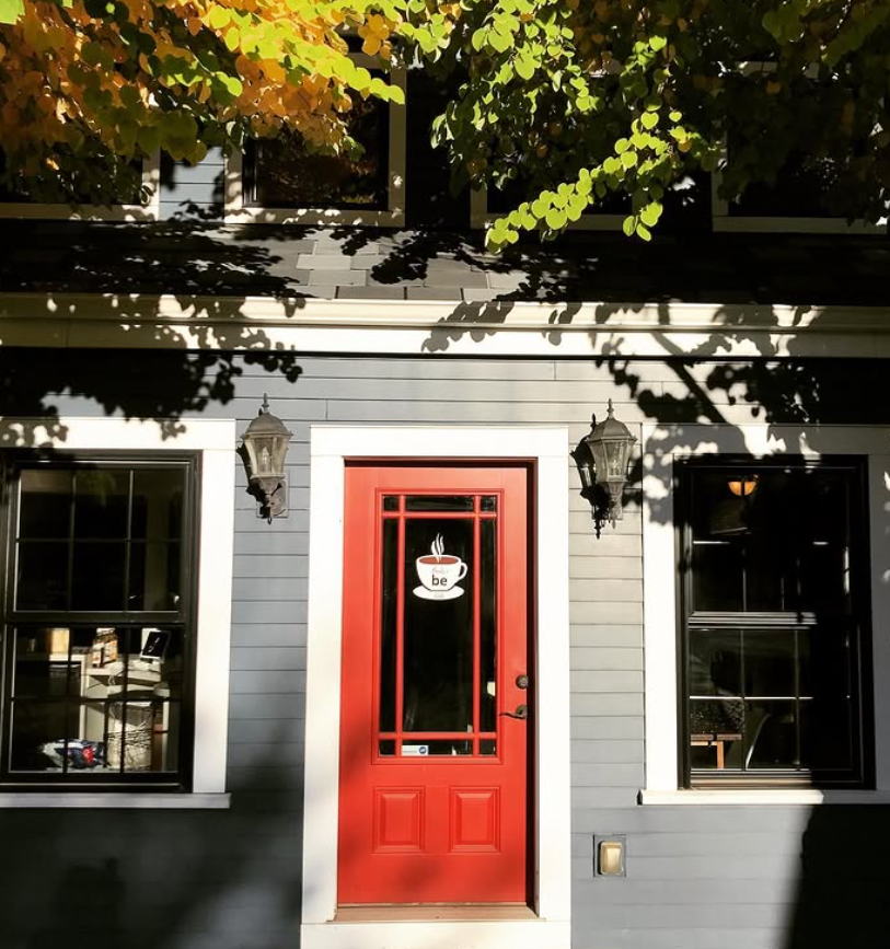 Front door of a house painted red with a window decal of a steaming coffee cup that says 'be'. There are two outdoor lanterns on either side of the door. The house is gray with white trim, and there are windows on either side of the door. Tree branches and leaves cast shadows on the house and door.