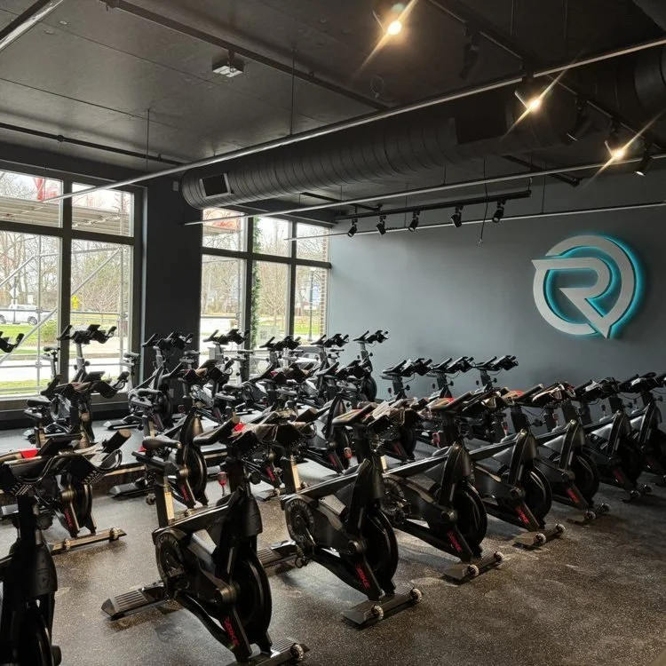 Indoor cycling class with multiple stationary bikes arranged in rows, a logo on the wall, large windows, and modern industrial design.