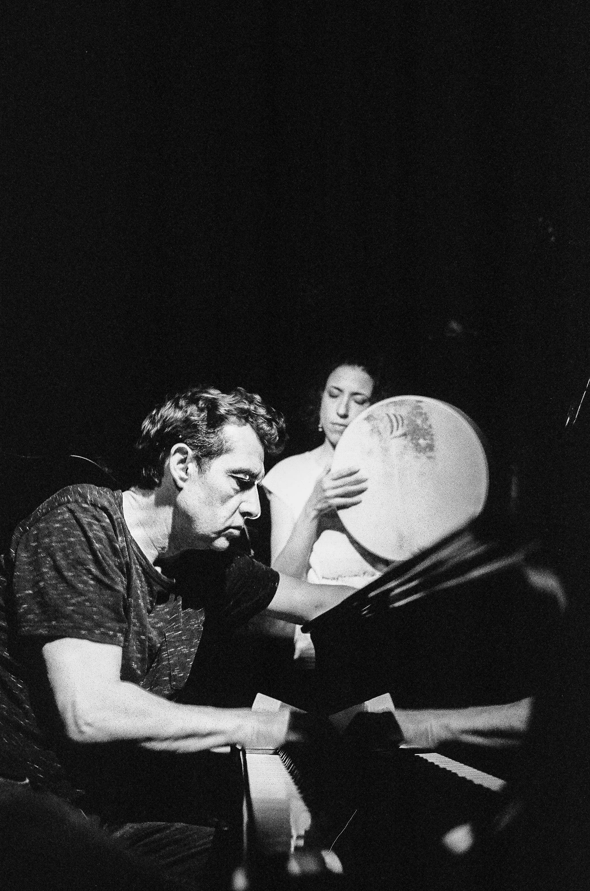 Deux musiciens, un homme au piano et une femme tenant un tambour, en activité de musique dans une scène monochrome.