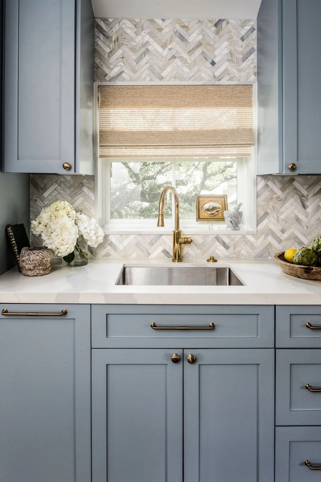 Kitchen sink with blue cabinets, a window with wood blinds, a gold faucet, a vase with white flowers, framed art, and a bowl of vegetables on the countertop.