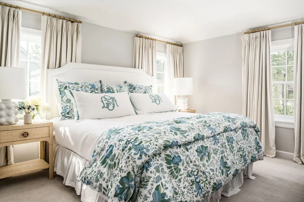 Bright bedroom with white walls, cream curtains, a white bed with floral bedding, decorative pillows with monograms, and nightstands with lamps and a flower vase.