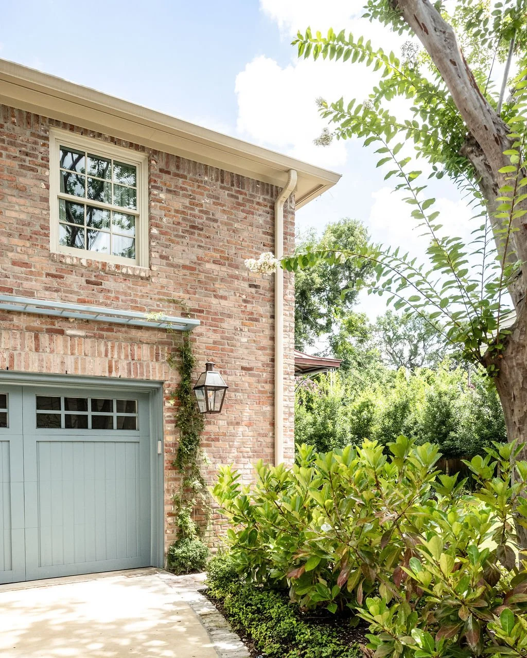 Exterior view of a brick house with a green garage door, a window above, a hanging lantern, and surrounding greenery including shrubs and trees.