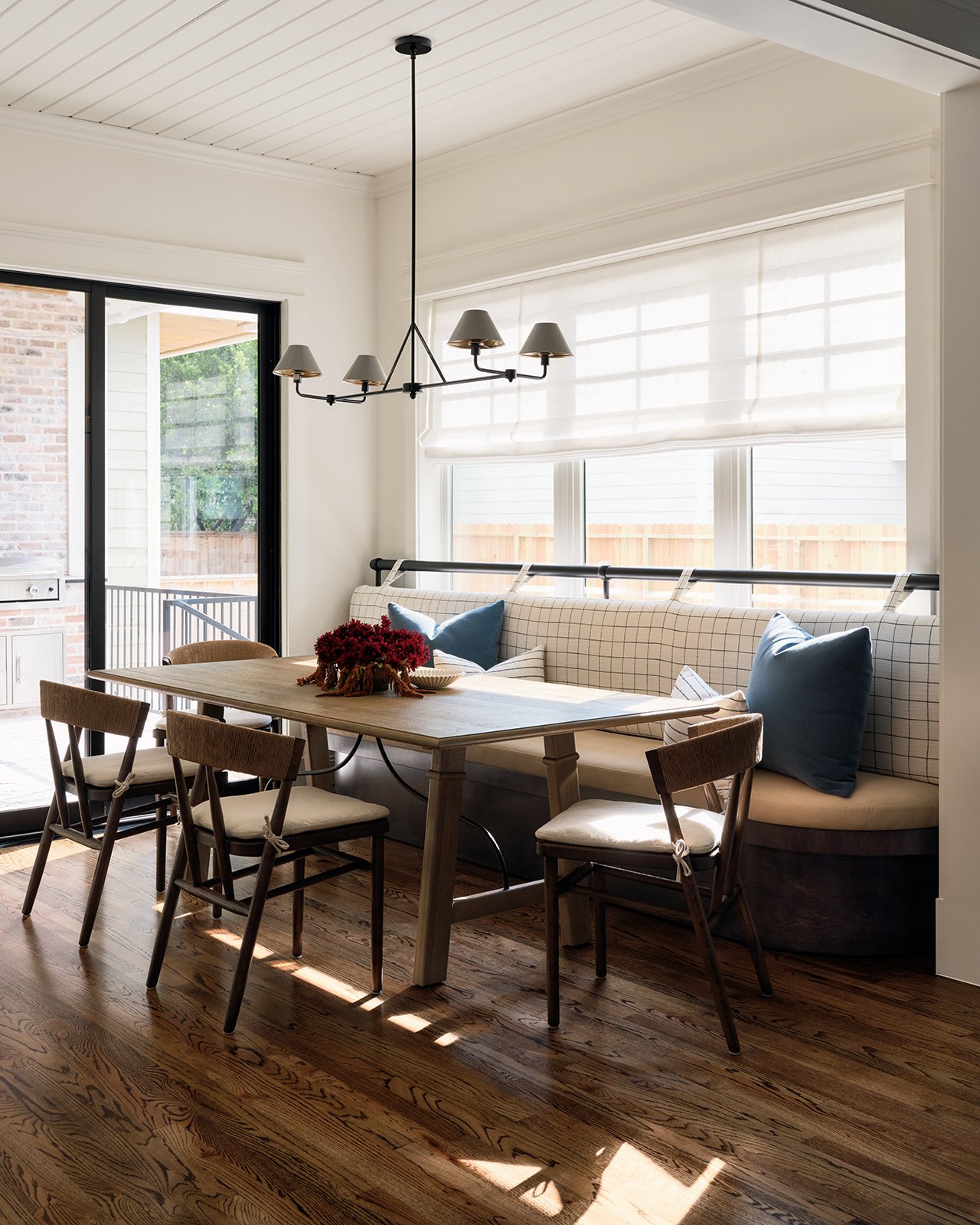 A dining area with a wooden table, four wooden chairs with white cushions, a built-in bench with checkered and solid blue pillows, and a chandelier hanging from the ceiling. The room has hardwood floors, large windows with white blinds, and a sliding