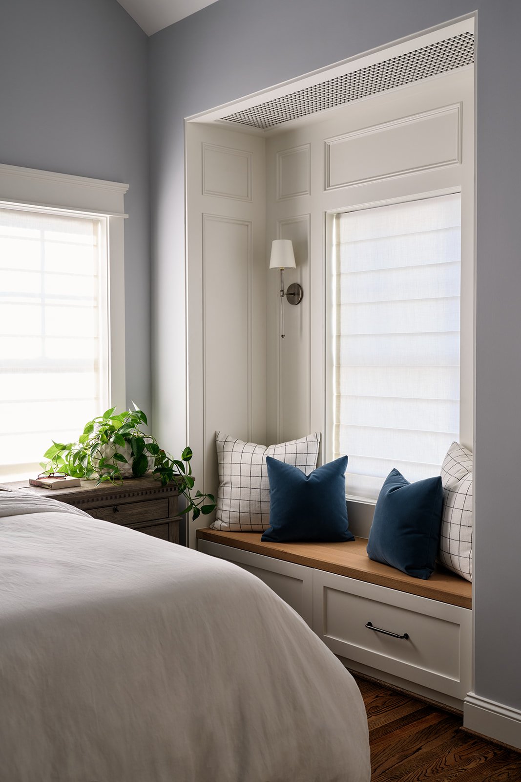 A cozy bedroom corner featuring a window seat with blue and plaid pillows, a built-in drawer, a window with white blinds, a wall sconce, and a window with a potted plant on a nightstand.