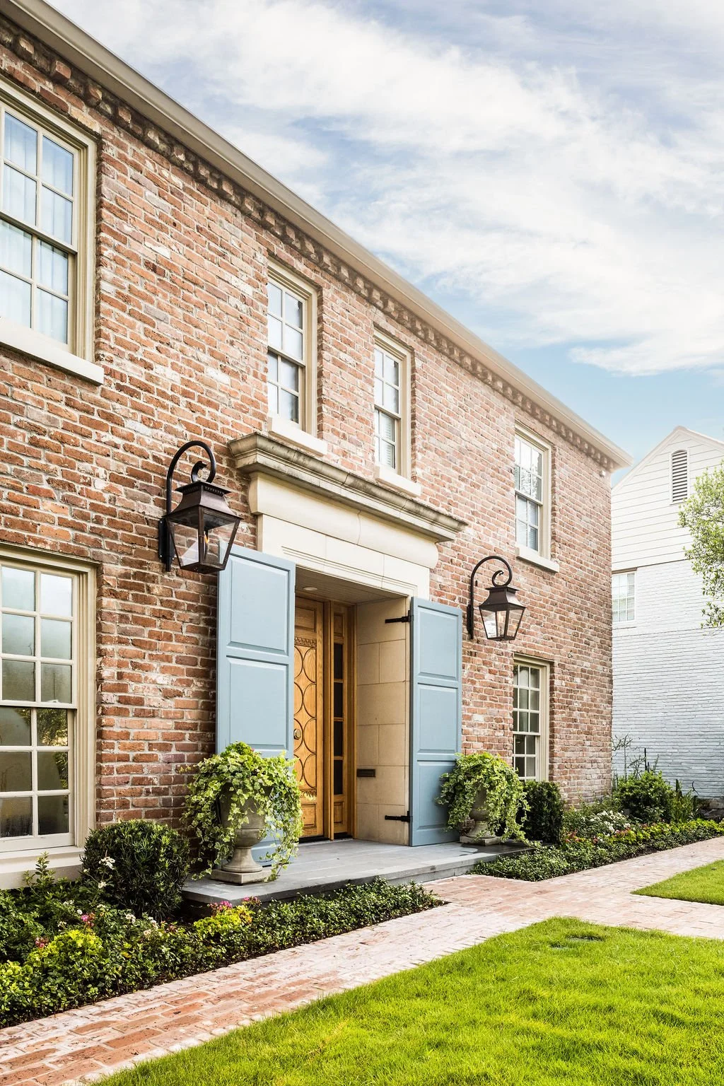A brick house with light blue shutters, a wooden front door, hanging lanterns, potted plants, and a well-maintained lawn.