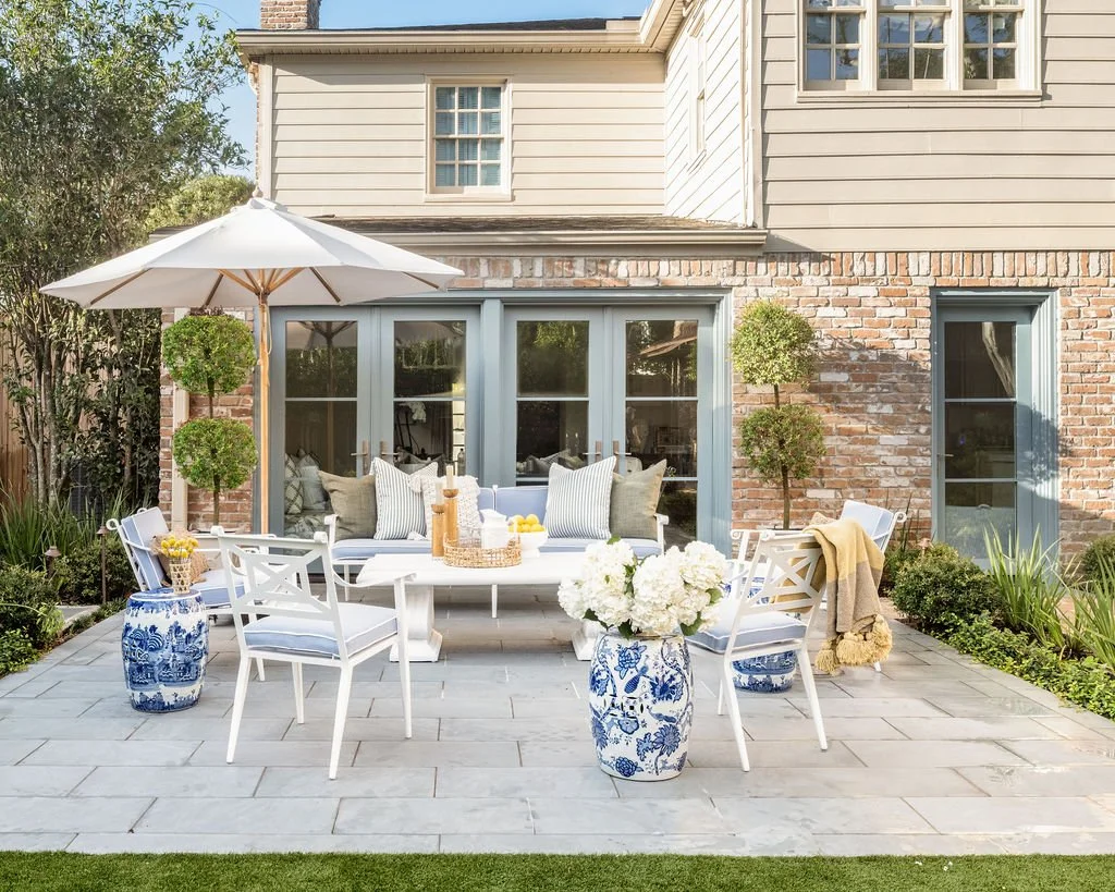 A cozy outdoor patio with a white table and chairs, large blue and white vases, an umbrella, and plants in front of a house with brick and siding exterior.