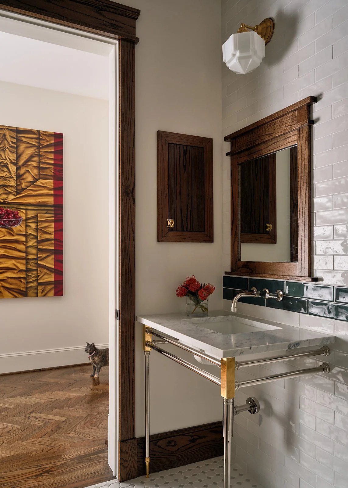 A bathroom corner with a marble sink, a mirror in a wooden frame, a small wooden cabinet, and a wall-mounted light fixture. A vase with red flowers is on the sink. A door frame shows a room with hardwood floors, a painting, and a gray tabby cat.