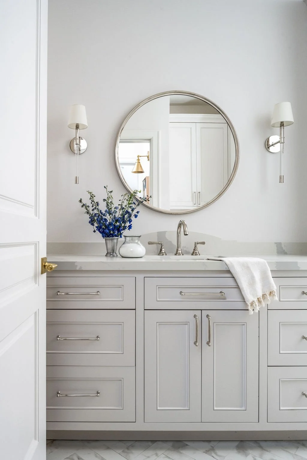 A white bathroom vanity with drawers and cabinets, a round mirror above, two wall-mounted lamps, a vase of blue flowers, a white soap dispenser, a silver faucet, and a towel on the counter.