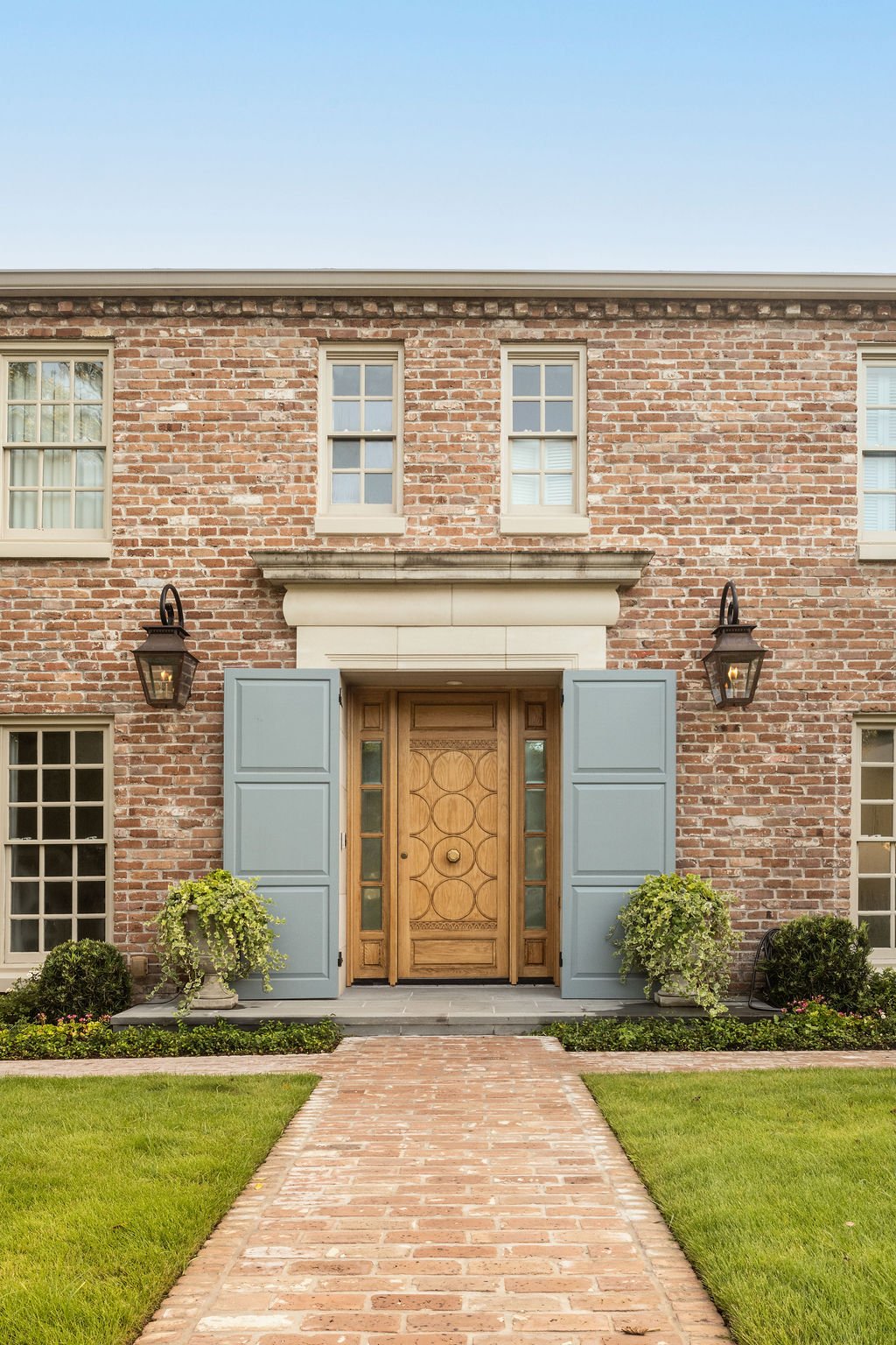 Classic River Oaks brick home renovated with a white oak front door, light blue shutters, and two Bevelo gas lanterns, with a brick walkway and green lawn.