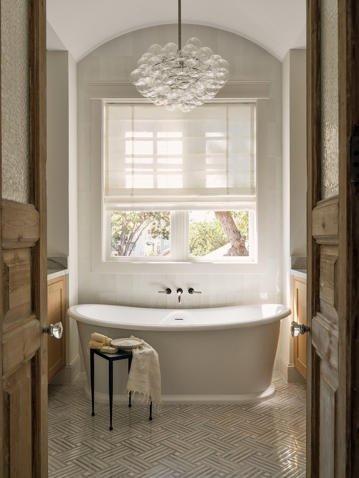 A bathroom with a white bathtub in front of a window with blinds, a black side table with towels and a soap dish, wooden cabinetry, and a glass bubble chandelier.