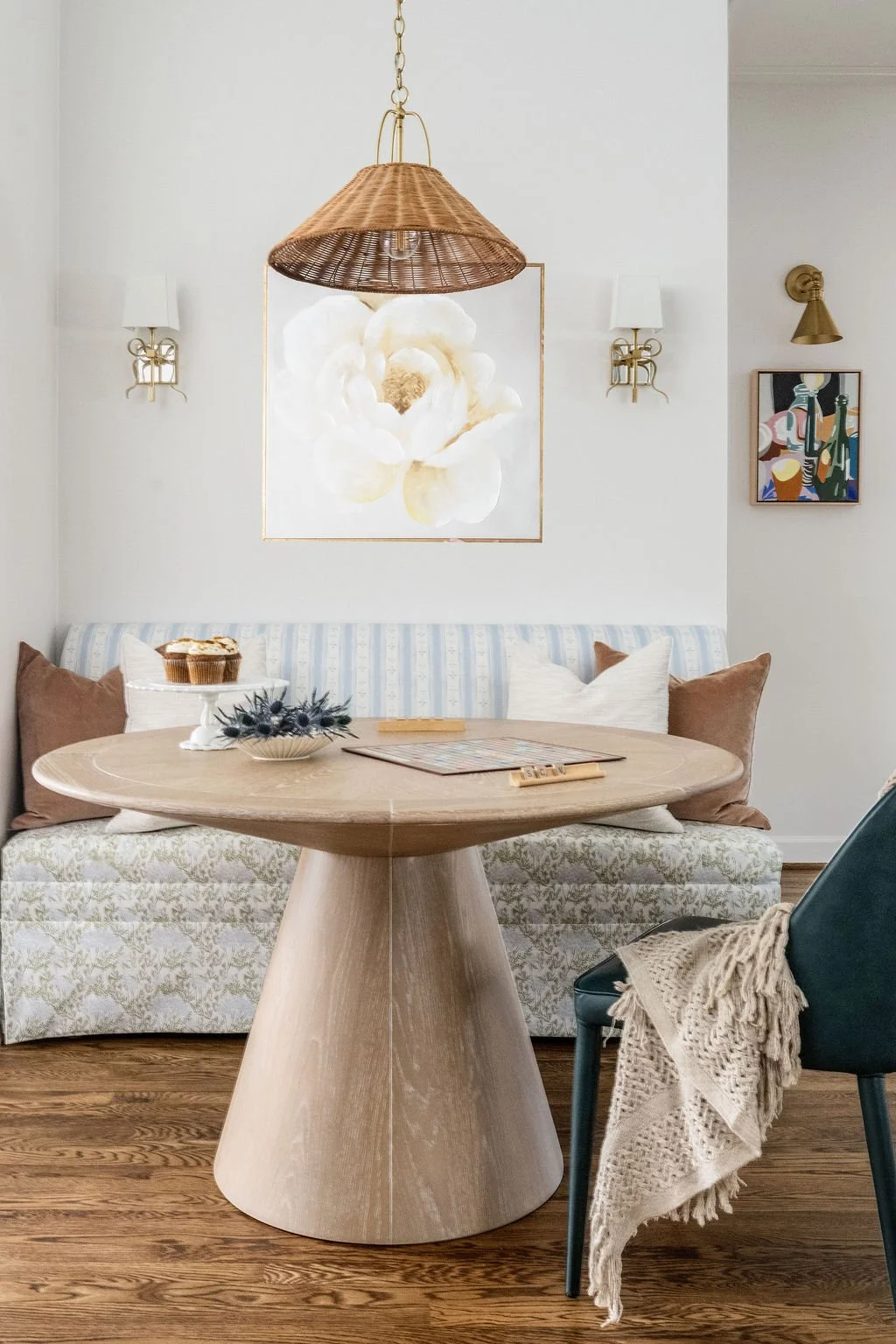 A cozy dining nook with a round wooden table, a striped cushioned bench, and a modern black chair. The table has a cake stand, a bowl with decorative objects, and a magazine. The wall behind features two white wall sconces, a large floral painting, a
