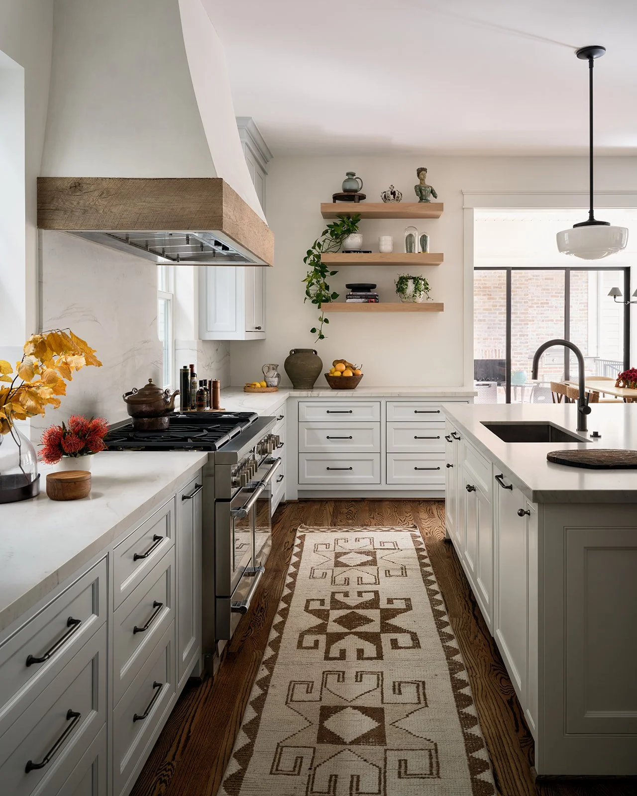 Modern white kitchen with wooden accents, open shelves, a patterned rug, and a view of a dining area with large windows.