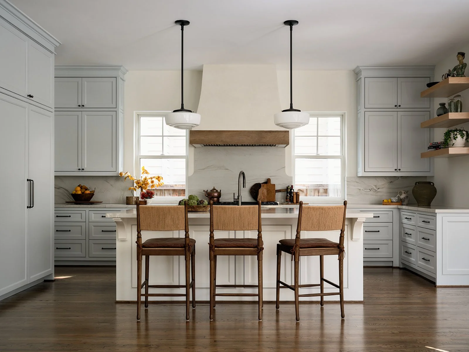 Modern kitchen with soft blue cabinets, marble backsplash, reclaimed beam, plaster venthood, wooden bar stools, and open wood shelves decorated with vases and books.