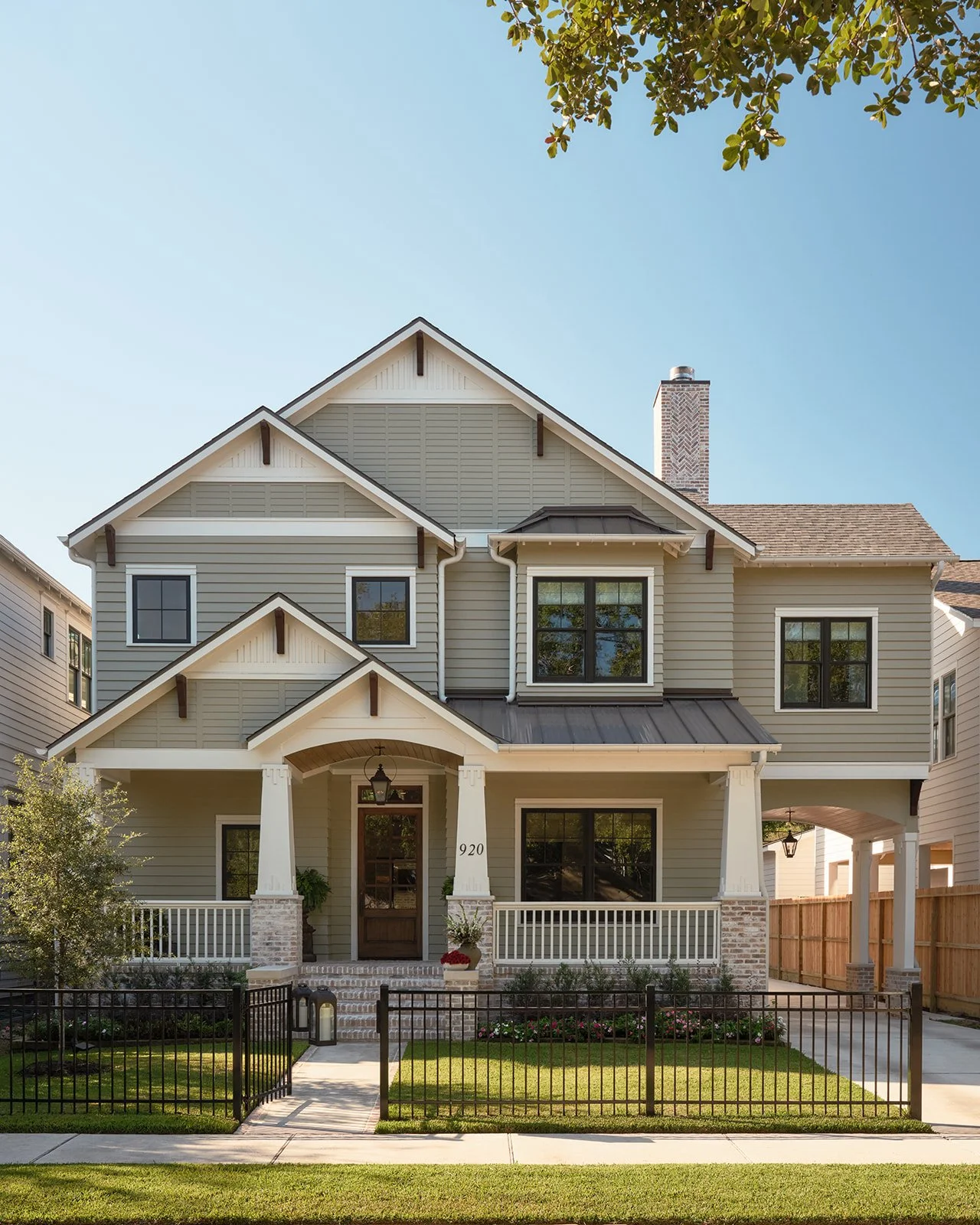 A modern, two-story house with beige siding, white trim, and a front porch with white railings. The house has multiple large windows, a gabled roof, and a brick chimney. There is a small, well-maintained front yard with a black metal fence, green gra