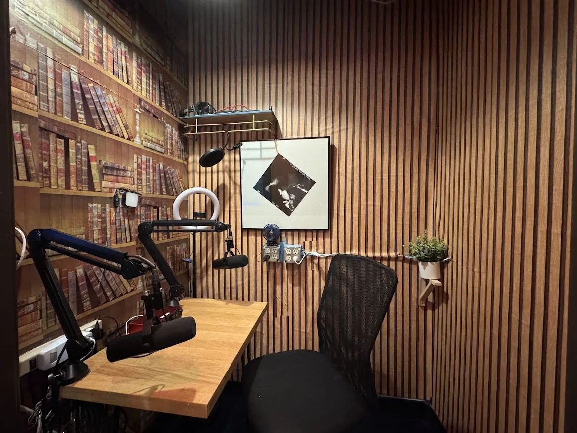 A cozy recording studio with wooden panel walls, a desk with microphones, headphones, a black mesh chair, a bookshelf with books, a framed photo on the wall, a shelf with headphones, a small plant on a wall-mounted shelf, and electrical outlets.